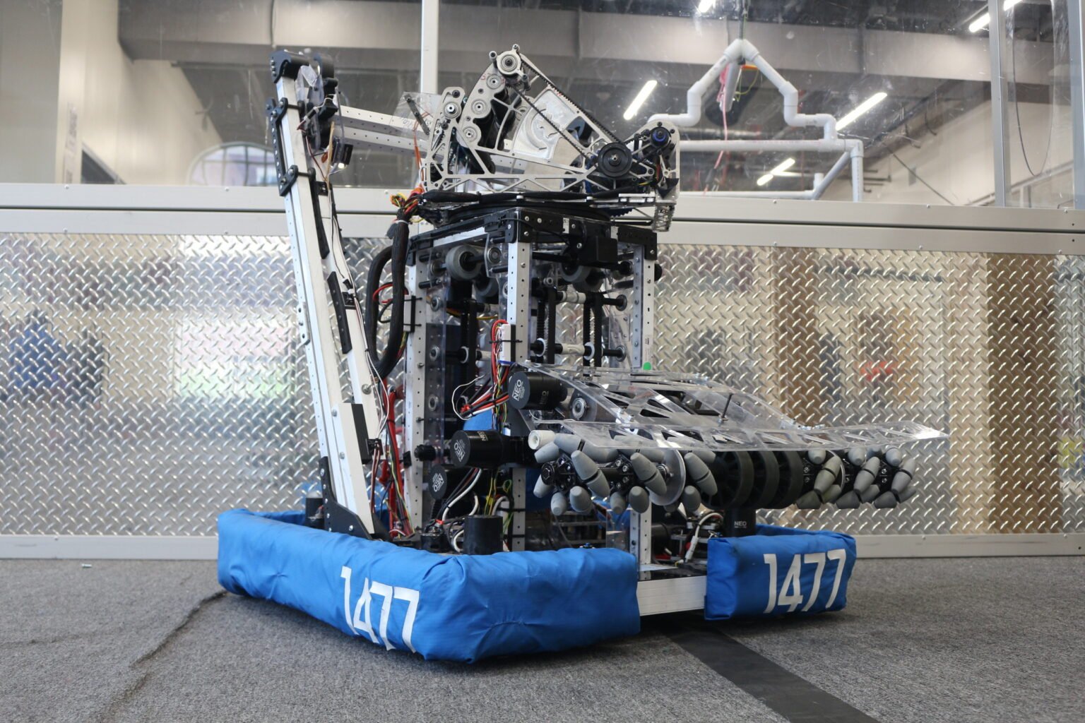 A robotics competition robot with the number 1477 on blue padded bumpers, featuring a mechanical arm and various electronic components, against a metallic wall and industrial background.