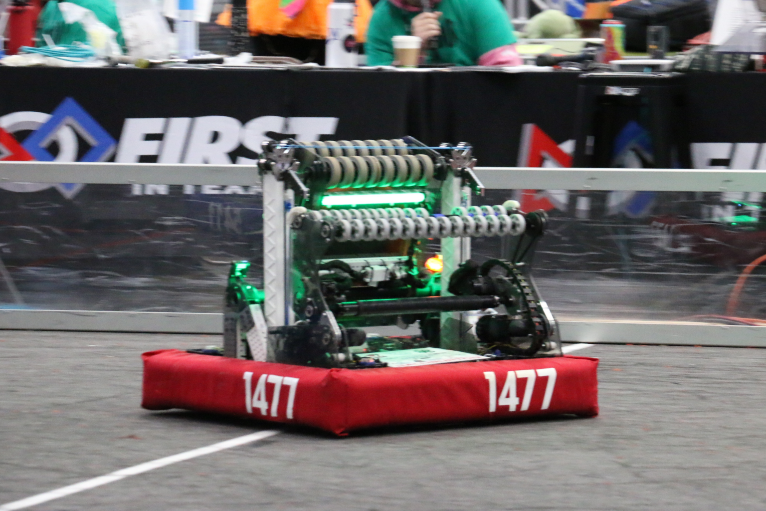 Competitive robot, "Takeoff", with the number 1477 on a red bumper, racing on a gymnasium floor at a robotics competition.