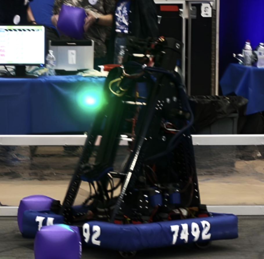 A robot on wheels with a green LED light, likely participating in a robotics competition, with people and equipment in the background.