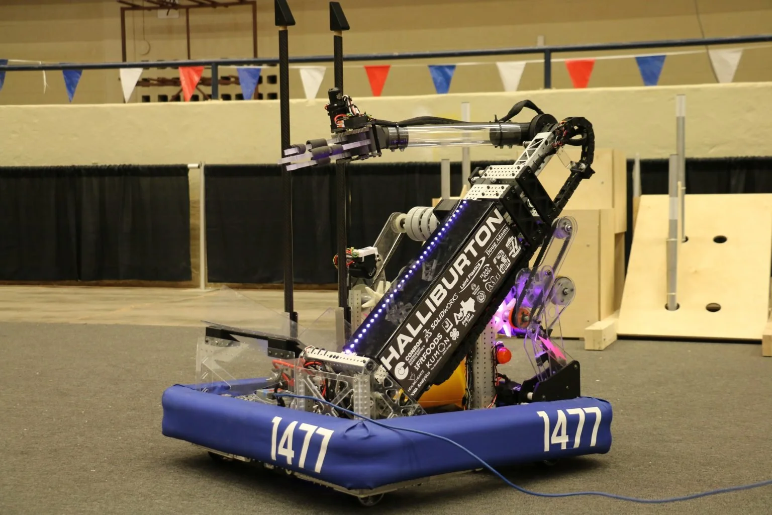 A robot with a black and blue color scheme, labeled with the number 1477, is in a competition setting with a wooden obstacle in the background. The robot features a long arm with sensors and mechanisms, and has LED lights on one of its panels.