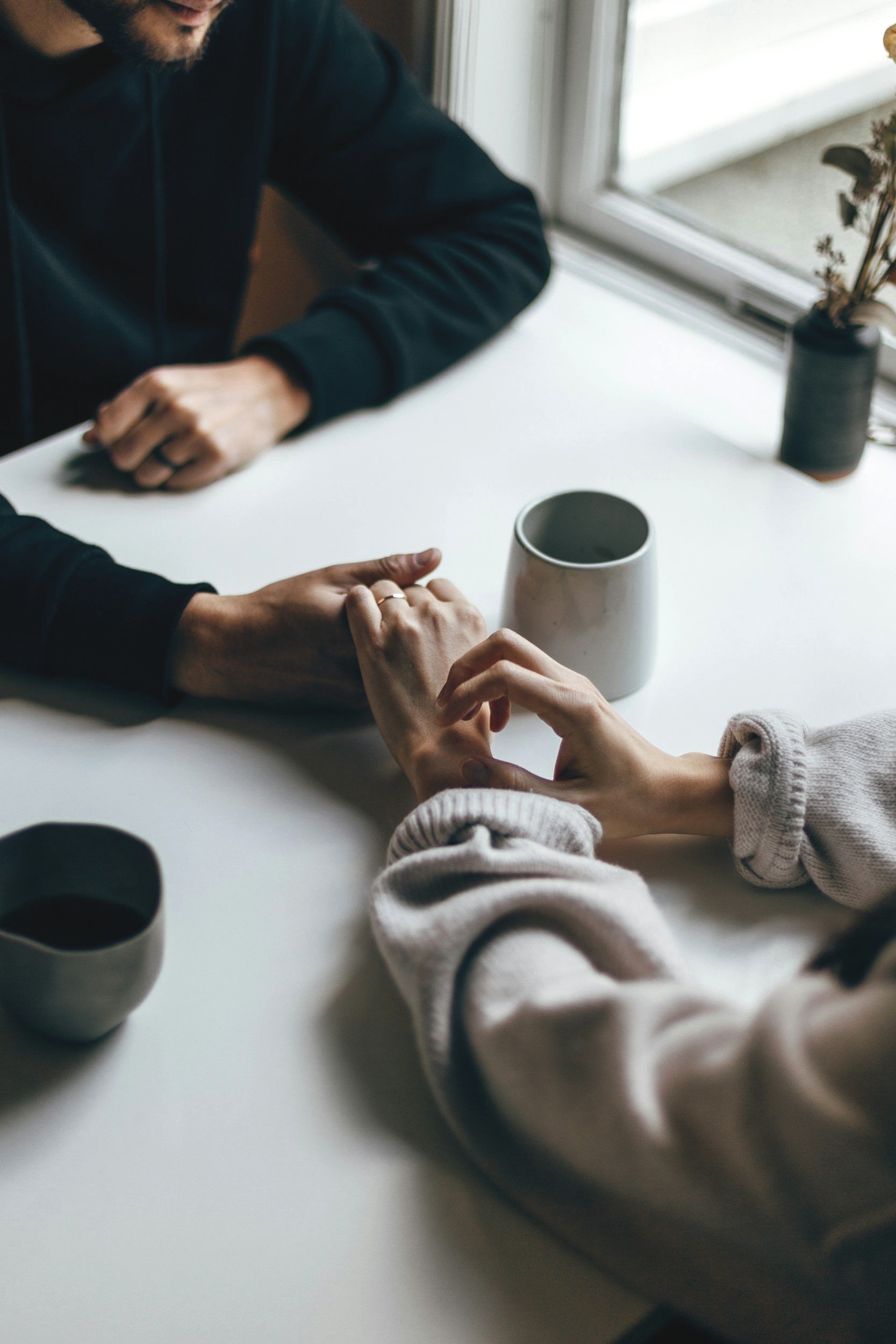 Two people holding hands across a white table near a window with two cups and a potted plant nearby.