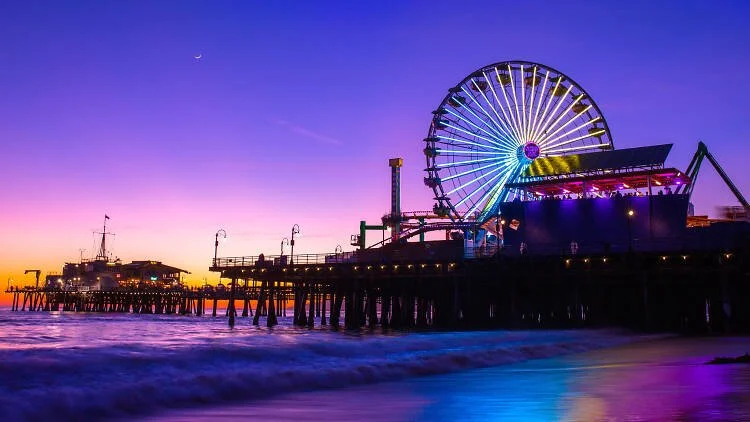 Colorful nighttime view of a pier with a lit Ferris wheel, amusement rides, and ocean in the foreground.