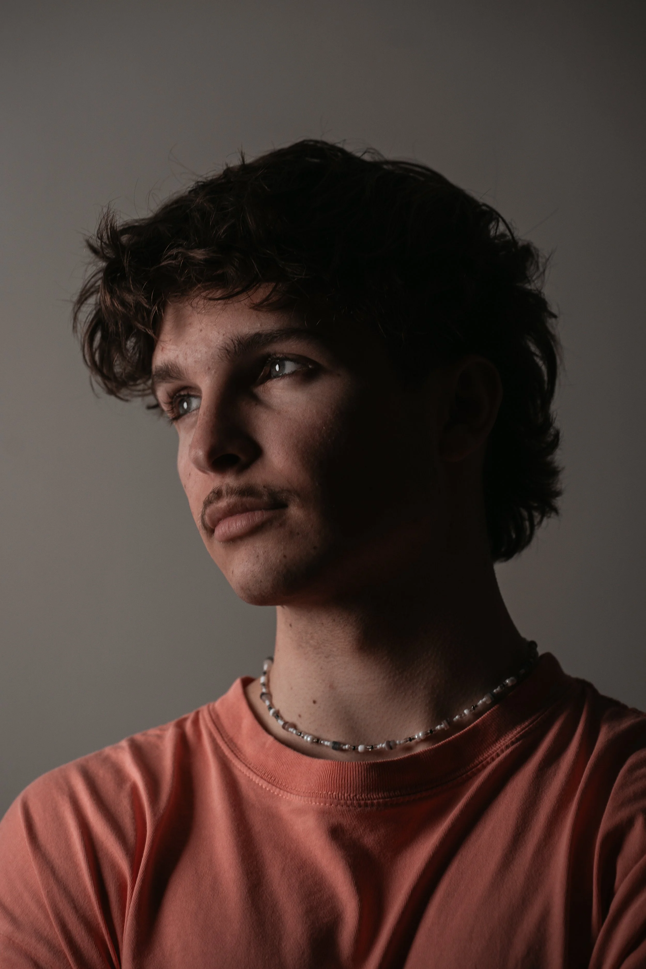 Porträt eines jungen Mannes mit braunen, lockigen Haaren, trinkt aus einer Tasse und blickt nach links, trägt ein pinkfarbenes T-Shirt und eine Perlenkette, vor grauem Hintergrund.