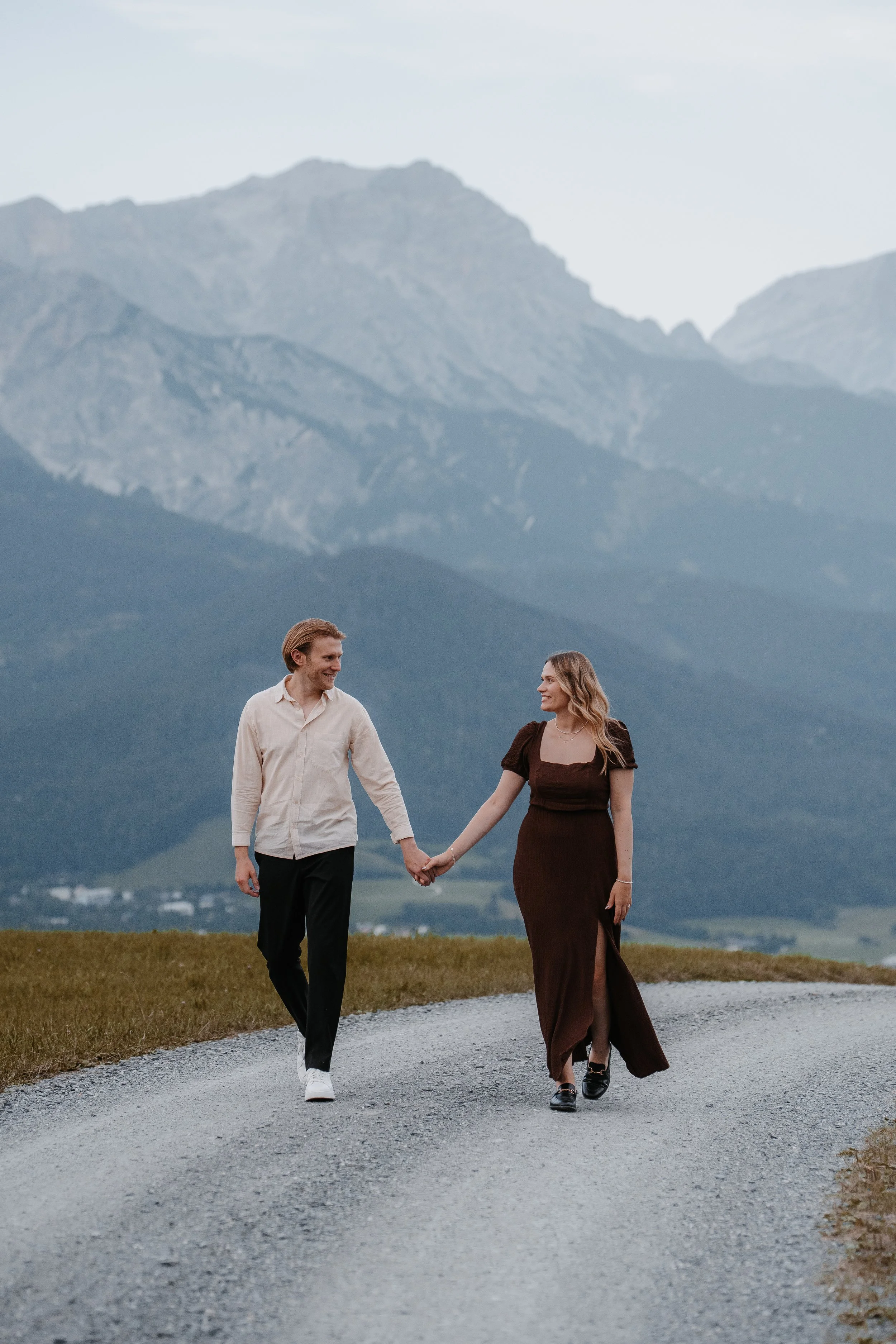 Ein Paar, das Hand in Hand auf einem Weg mit Bergen im Hintergrund spaziert.
