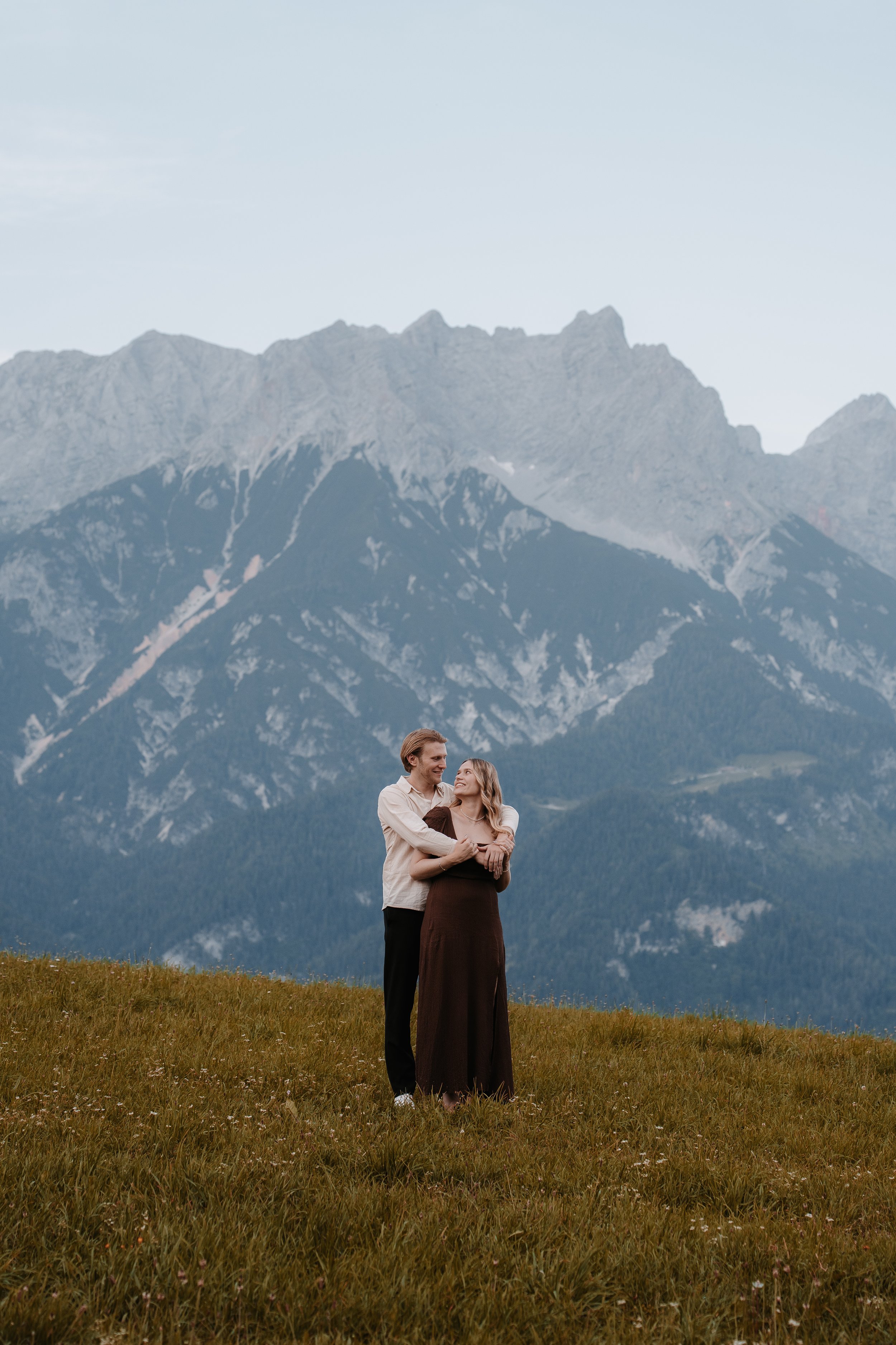 Ein Paar steht auf einer Wiese vor einer Bergkulisse und umarmt sich, während sie sich ansehen.