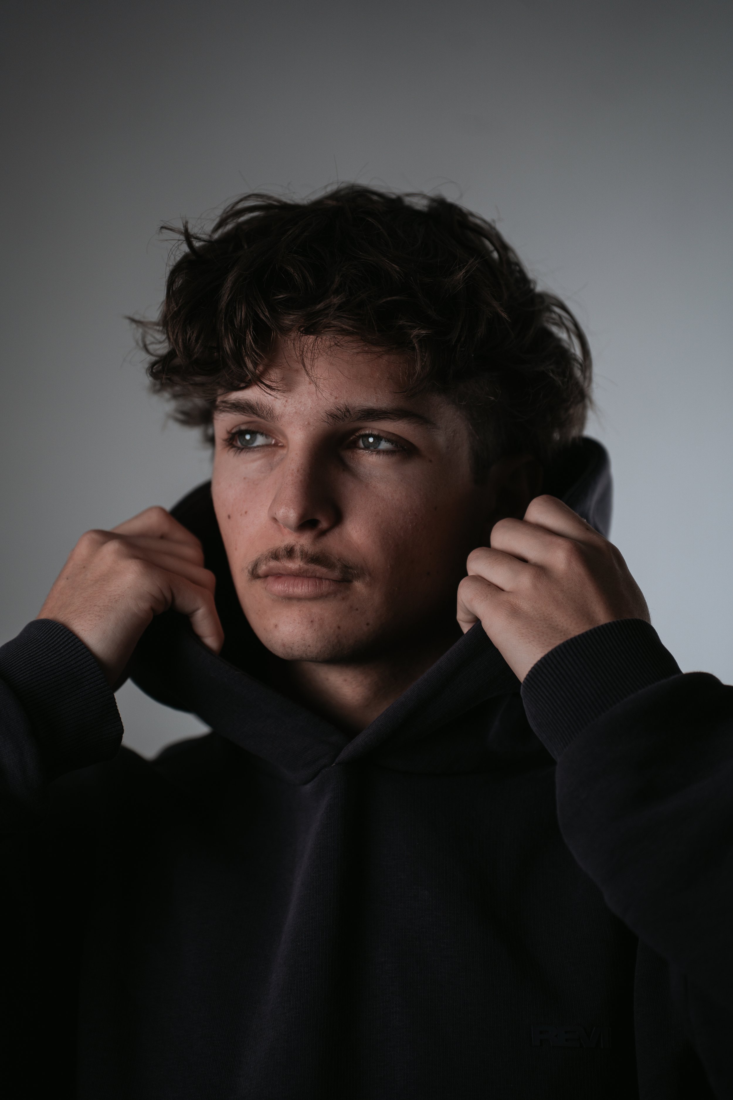 Junger Mann mit dunklen, lockigen Haaren trägt einen schwarzen Hoodie, schaut nach rechts, hält den Hoodie mit beiden Händen am Hals.