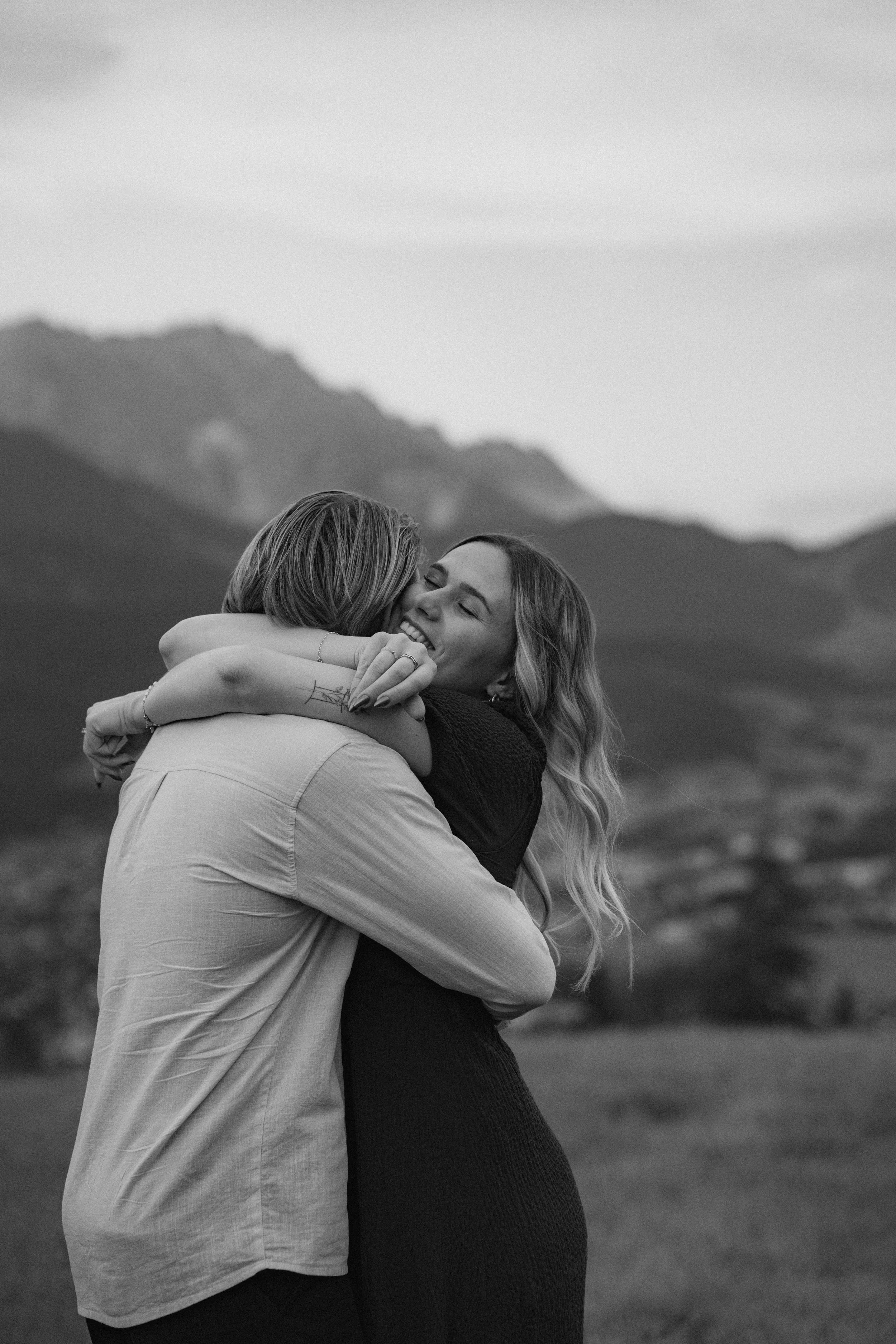 Zwei Frauen umarmen sich liebevoll in einer natürlichen Landschaft mit Bergen im Hintergrund, beide lächeln glücklich.