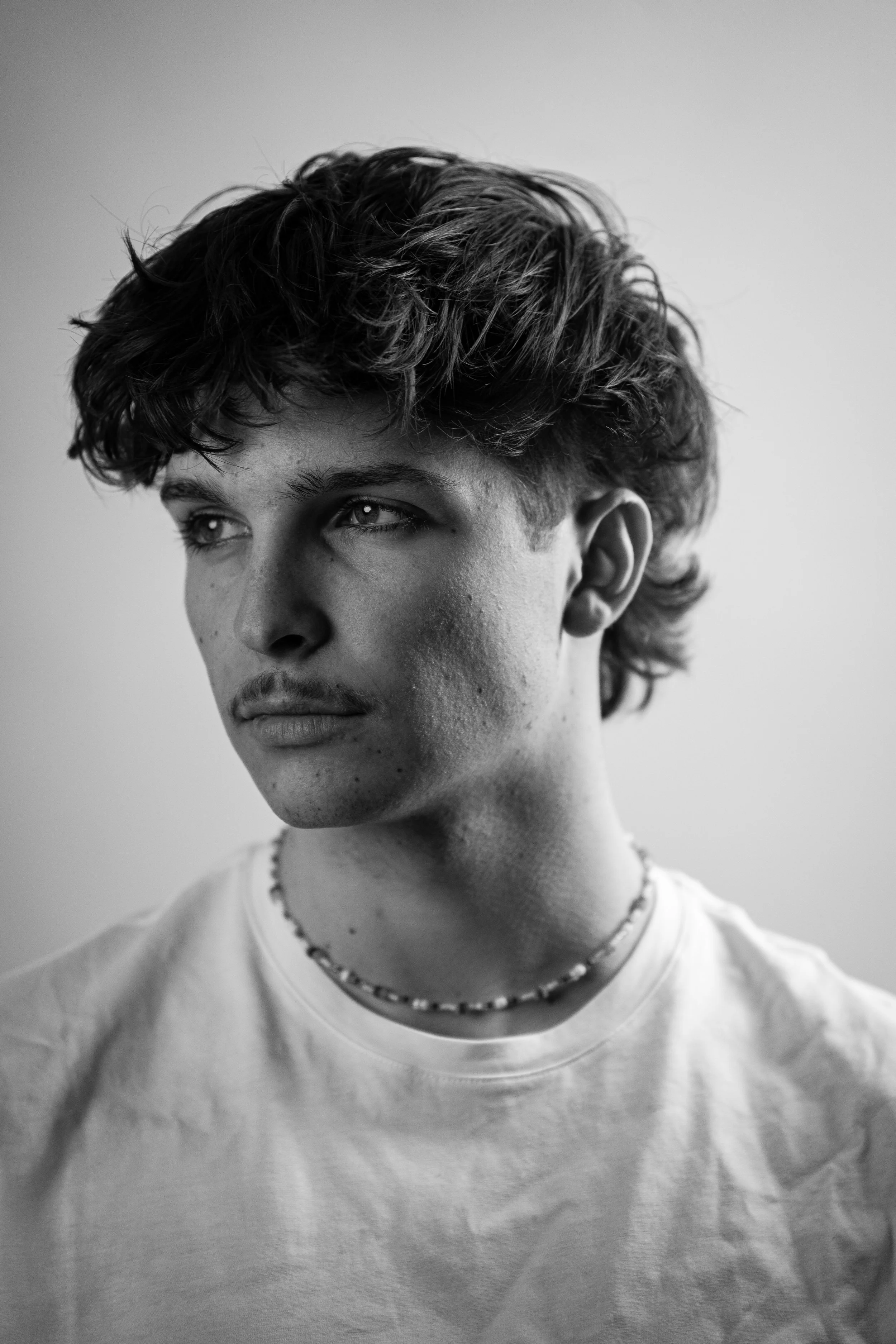 Schwarz-weiß-Porträt eines jungen Mannes mit lockigem Haar, ein Blick nach rechts, wearing a T-Shirt and einer Perlenkette.