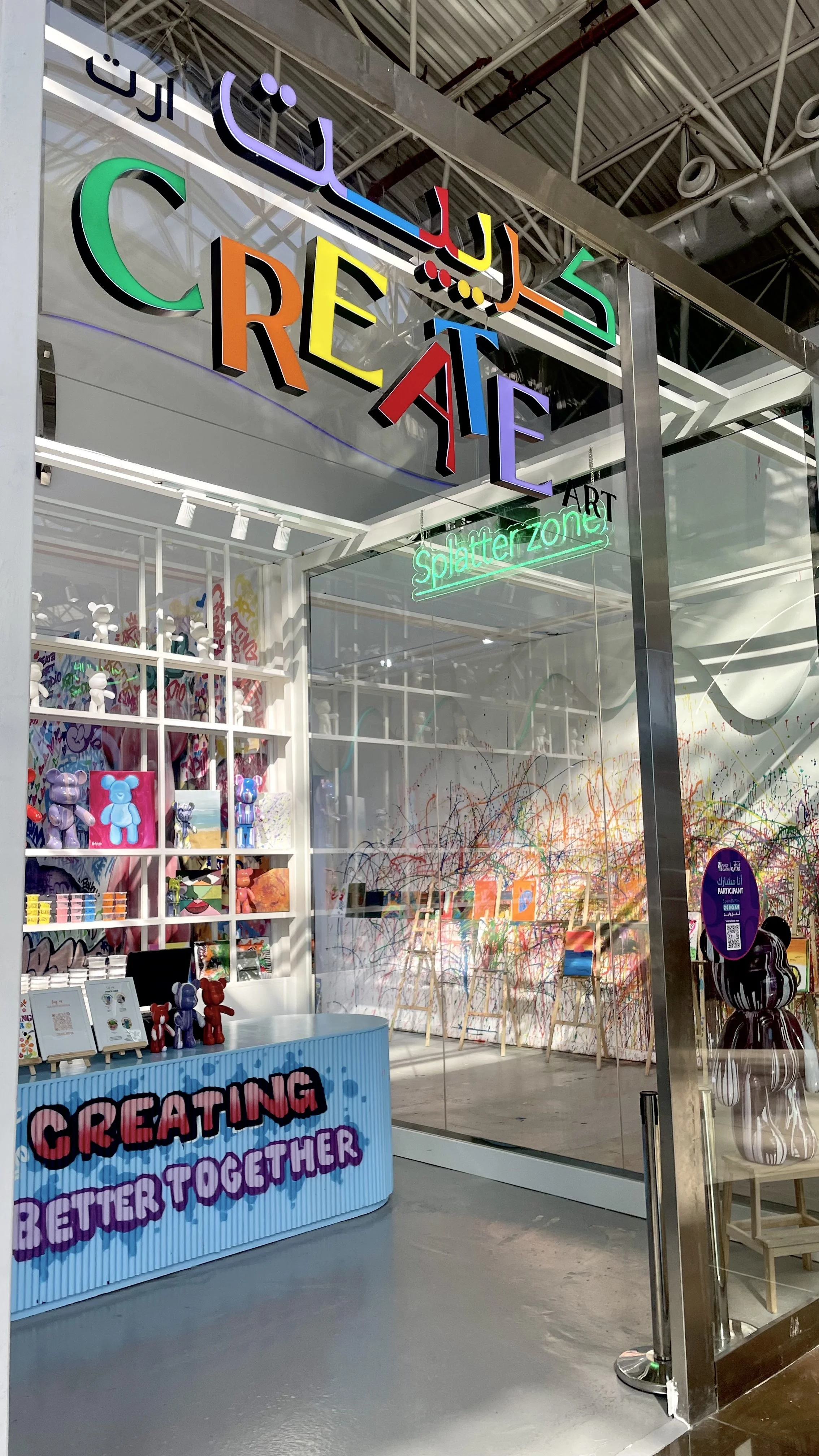 A colorful art shop with a sign that reads 'Create' in large multi-colored letters, next to a neon sign that says 'Splatter zone'. Inside, there are paint splatters on the wall, easels, shelves with art supplies, and a table with several art toys and
