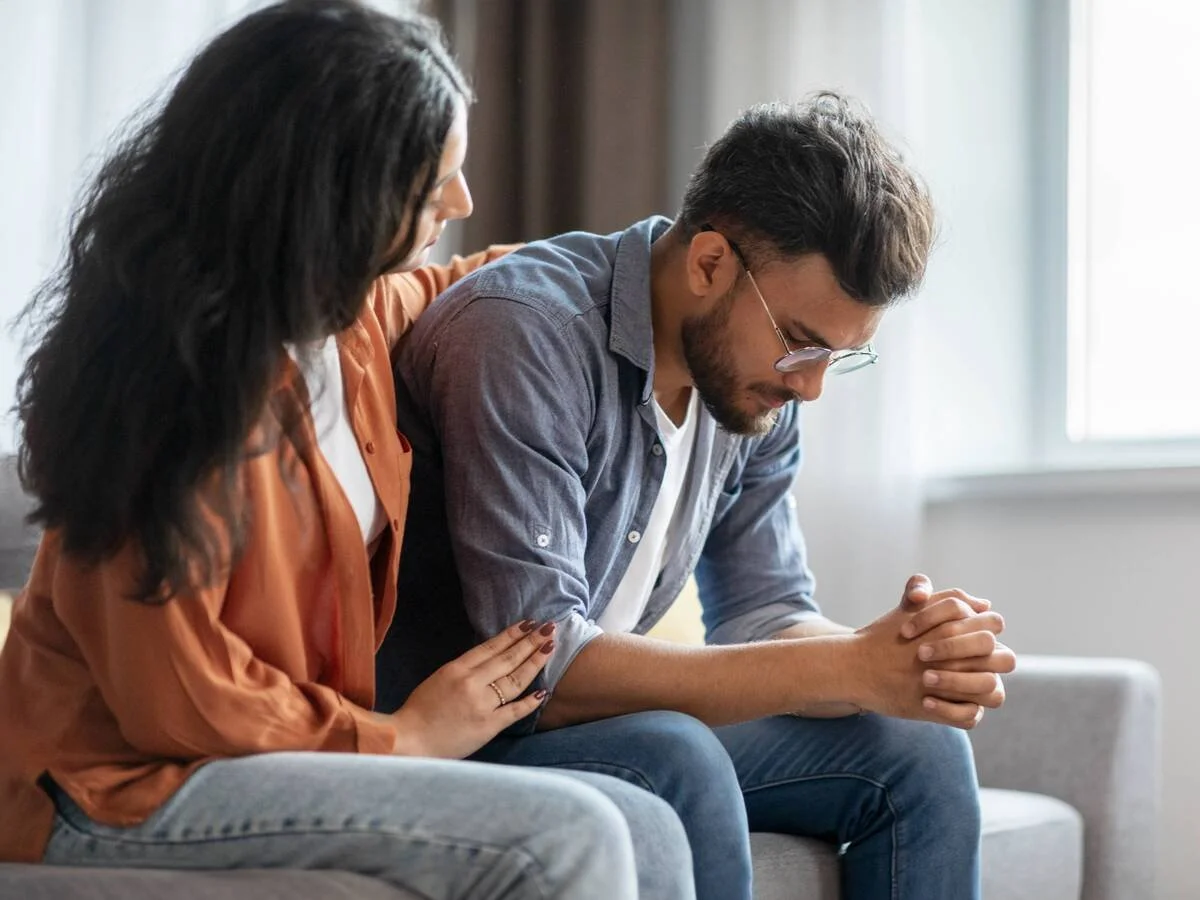 Woman gently reaching toward her partner while he withdraws and looks down, showing the shut-down and get-louder cycle in relationships.