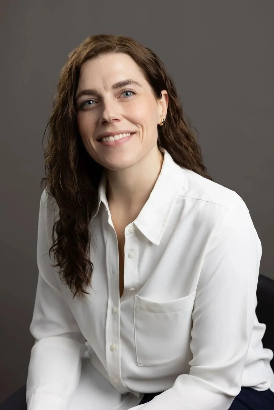 A professional photo of Andrea in a collared shirt with a warm smile, leaning forward seated on a stool.