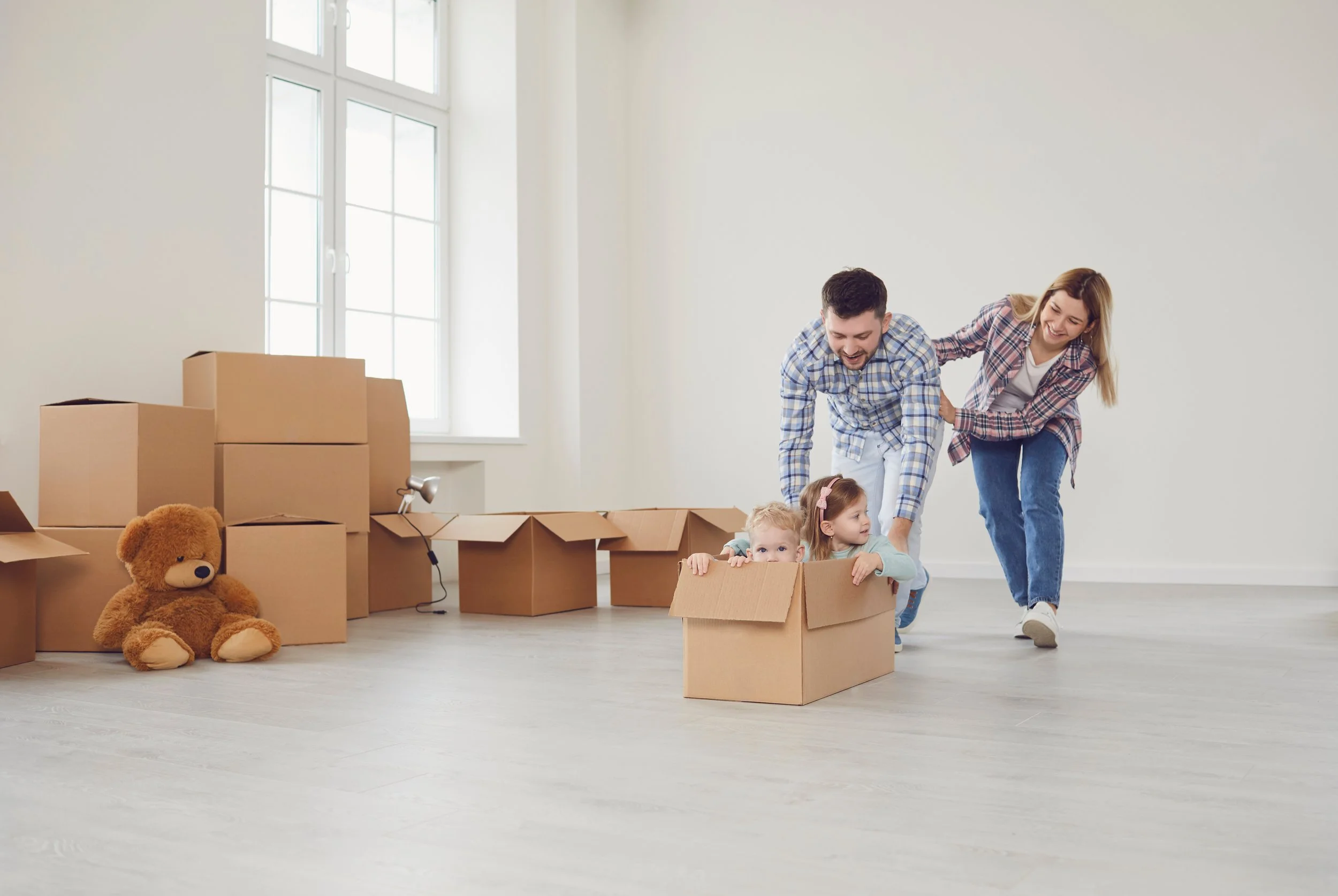 A family of four with two young children playing in an empty room filled with moving boxes, with one child sitting in a cardboard box and the parents helping to push them along.