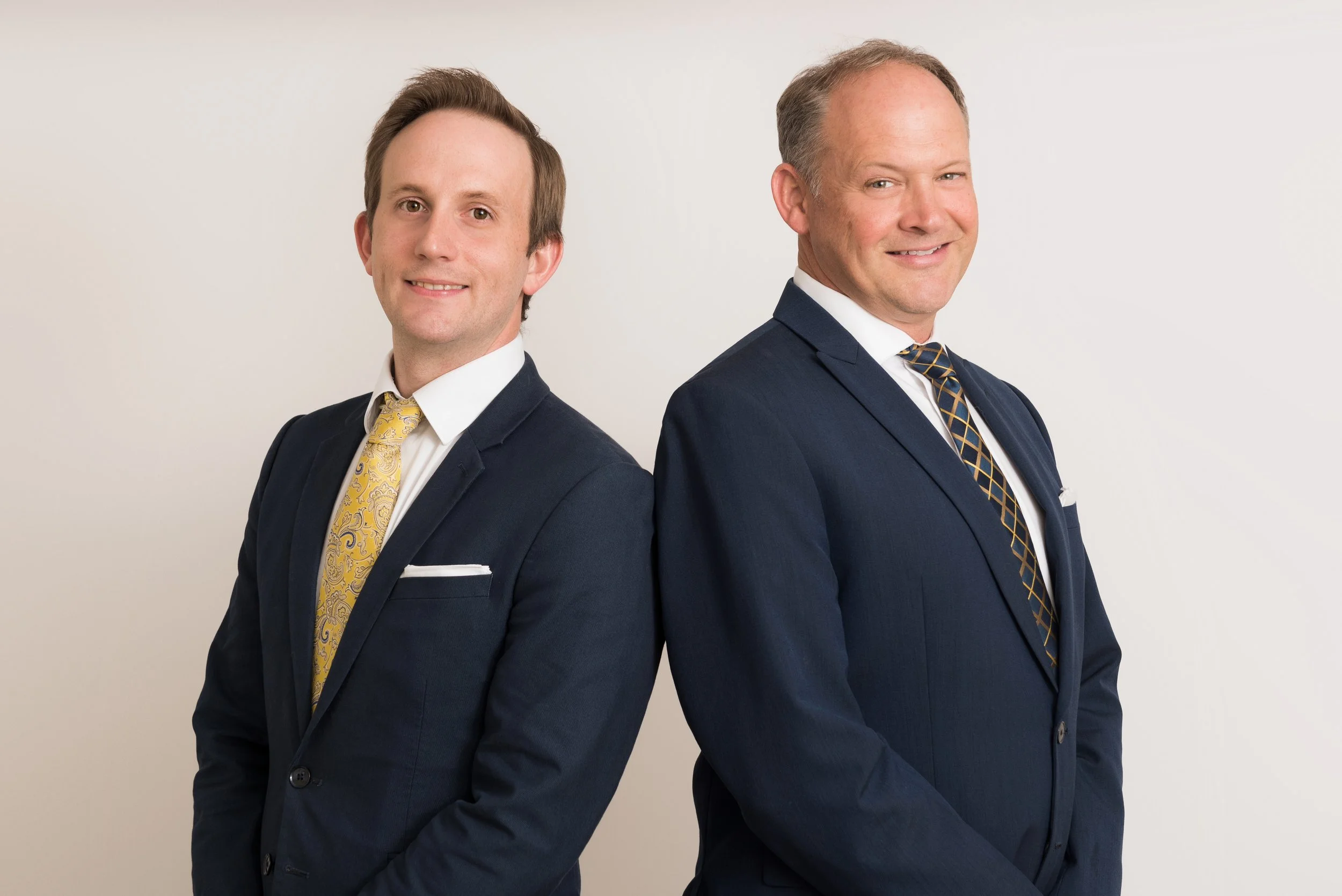 Two men in suits standing back-to-back, smiling, wearing navy blazers, white shirts, and patterned ties against a plain white background.