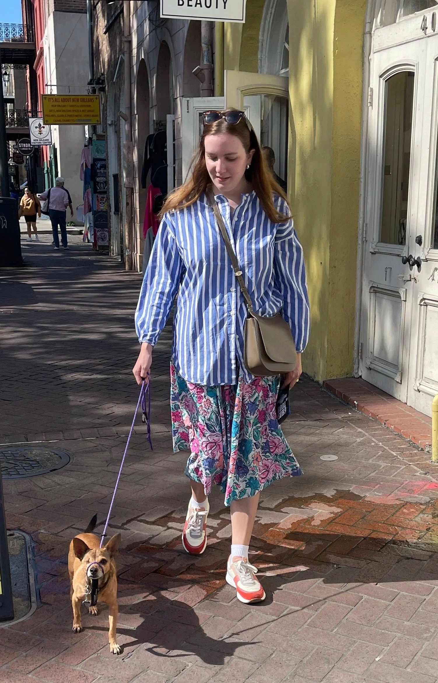 A woman walking her small brown dog on a city sidewalk. The woman is wearing a blue and white striped shirt, a floral skirt, and sneakers, with sunglasses on her head. The street has colorful buildings and signs in the background.