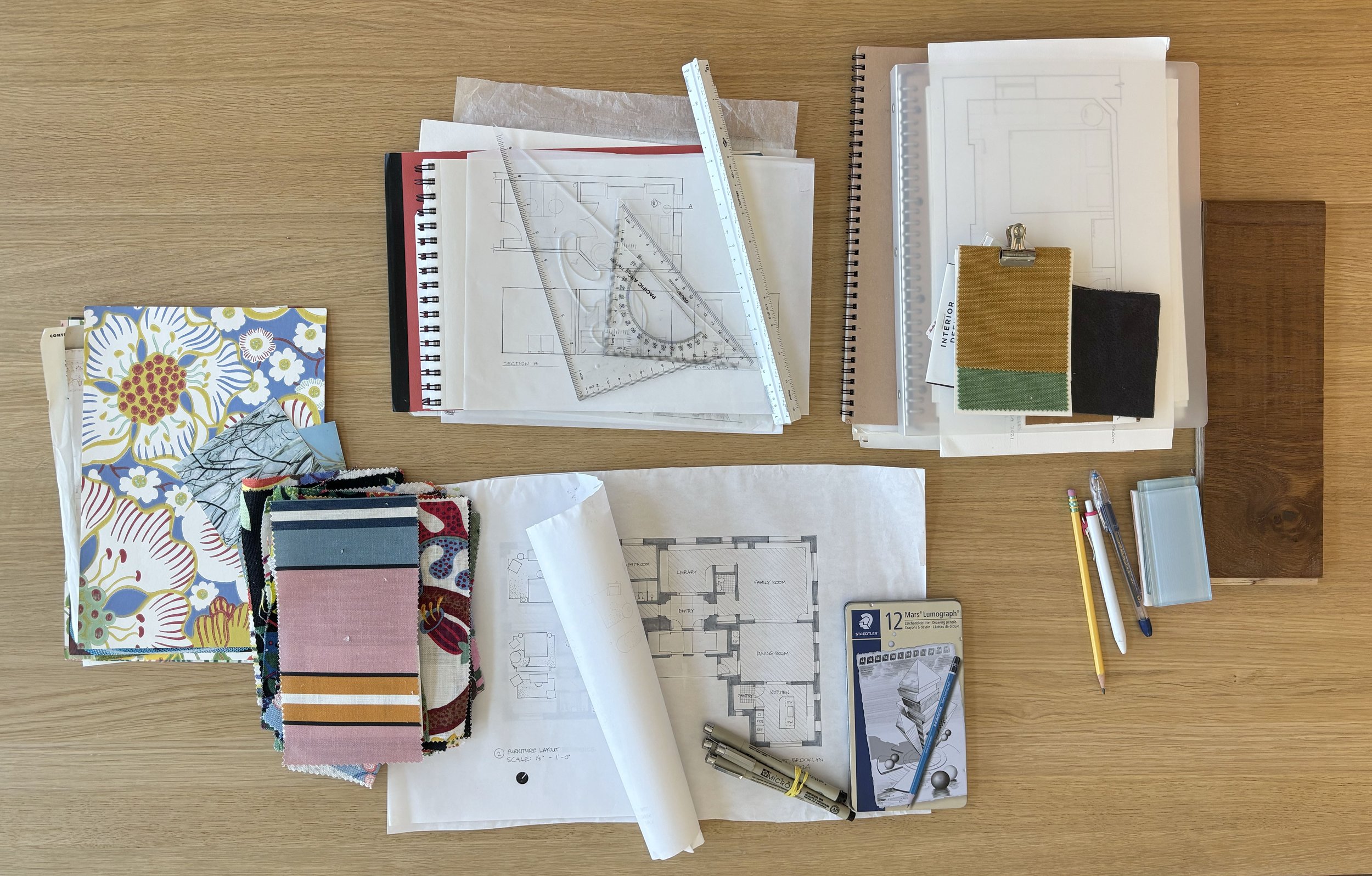 Architectural blueprints, fabric swatches, pens, pencils, rulers, and drafting tools on a wooden desk.