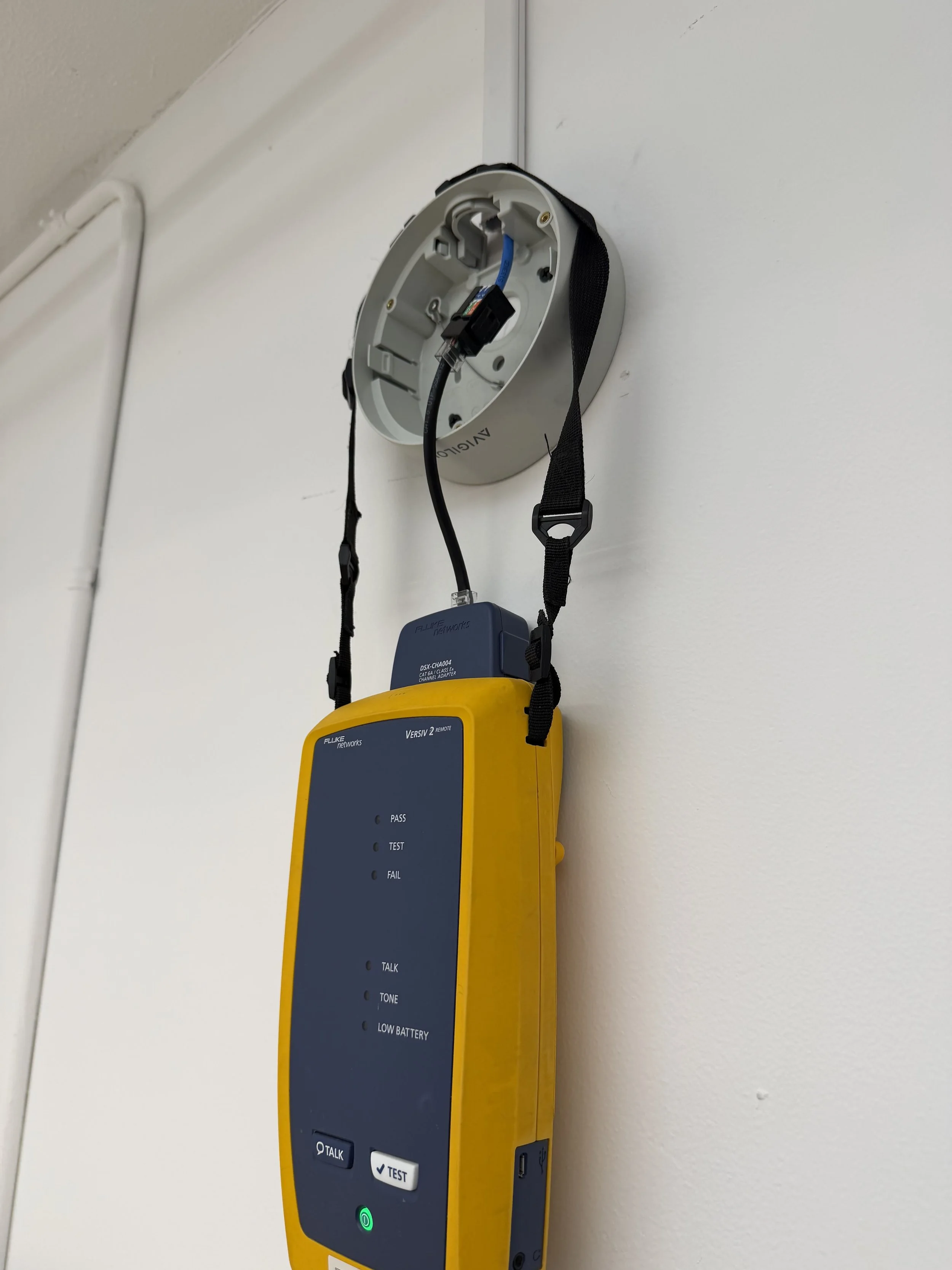 Cable tester connected to cabling at a security camera mounting point on the office wall.
