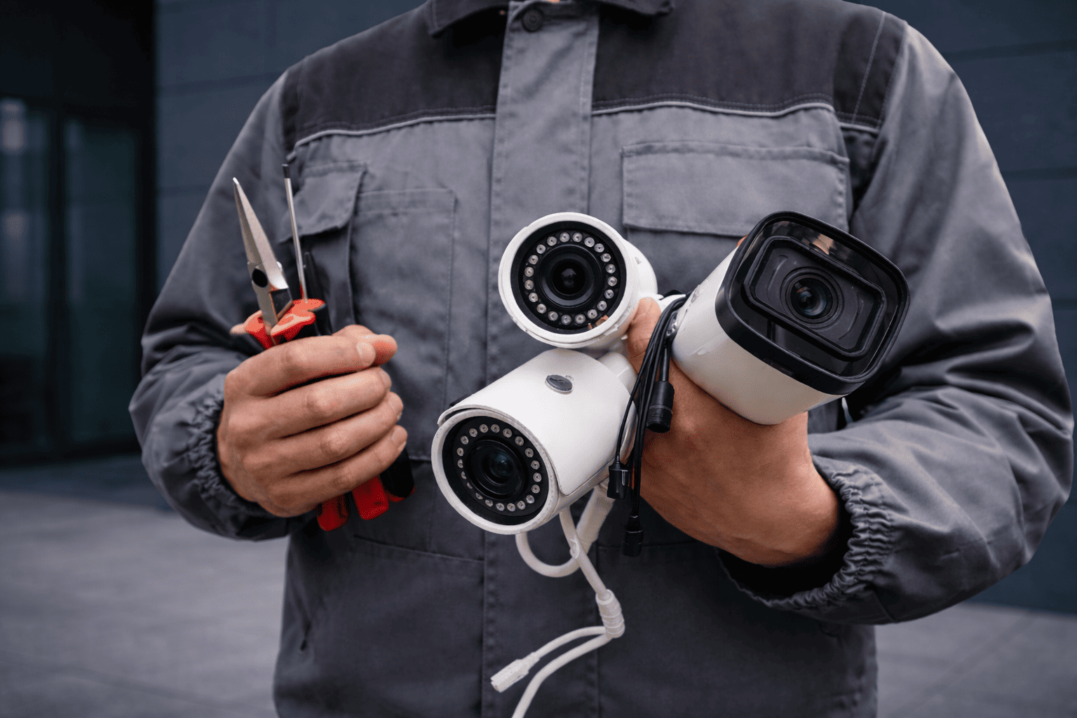 Security technician holding multiple commercial-grade bullet surveillance cameras and installation tools outside a modern building.