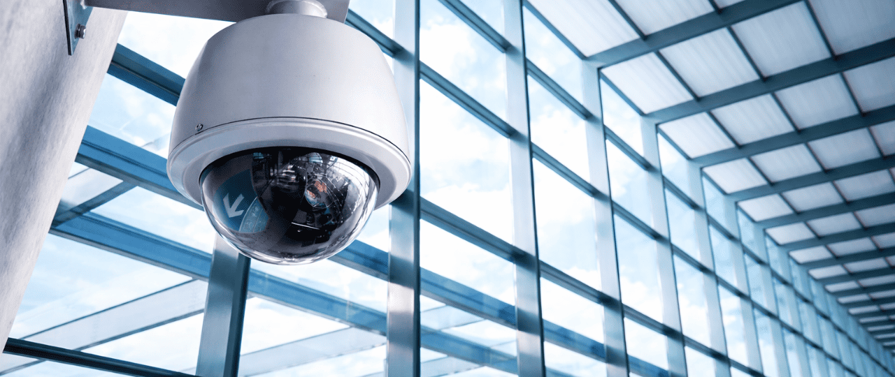 Dome security camera mounted indoors overlooking a modern commercial glass building interior.