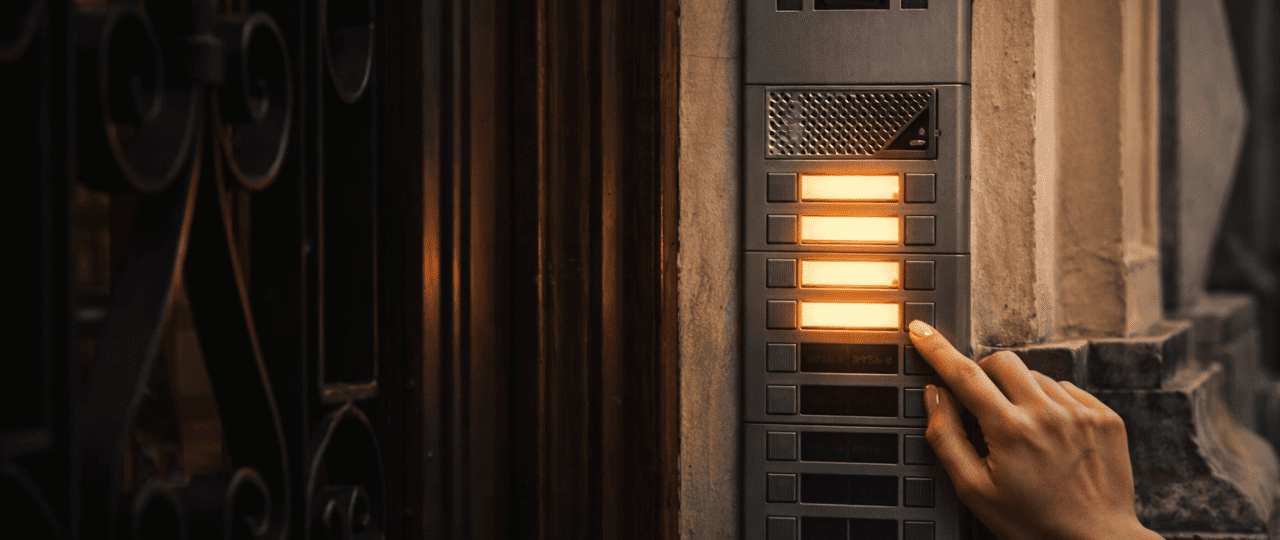 Resident pressing an illuminated apartment intercom button at a New York City building entrance, using a secure multi-tenant door entry system with backlit call buttons in an urban residential setting.png