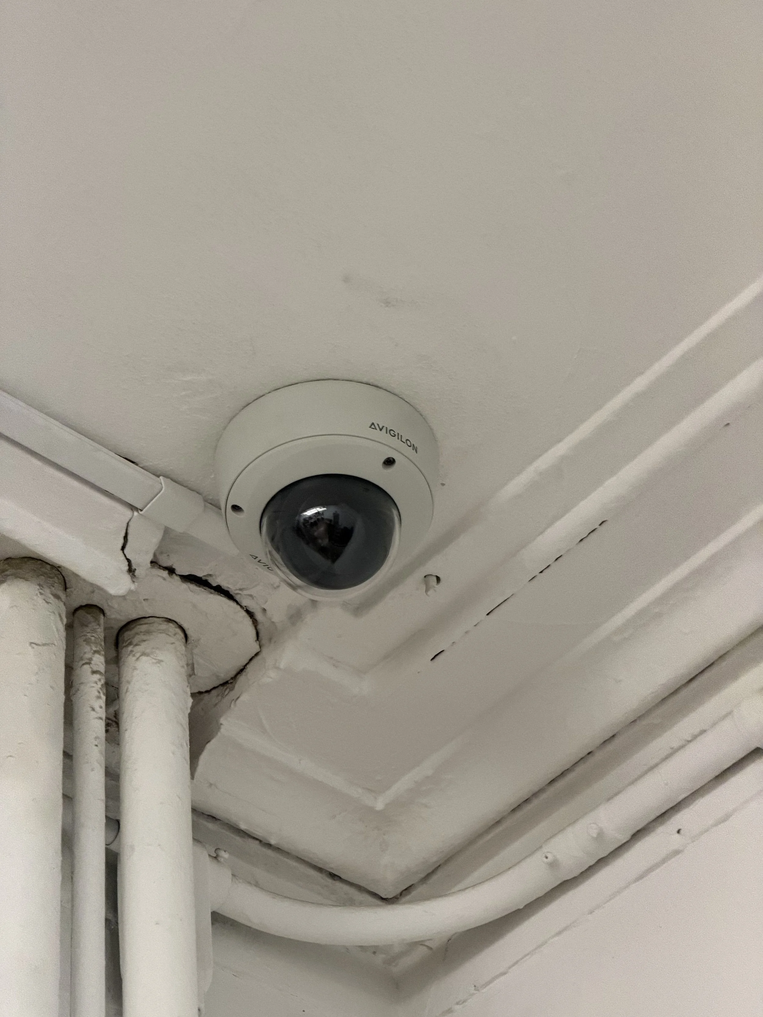 Dome security camera mounted near the ceiling beside exposed pipes, with white surface raceway carrying its cable.