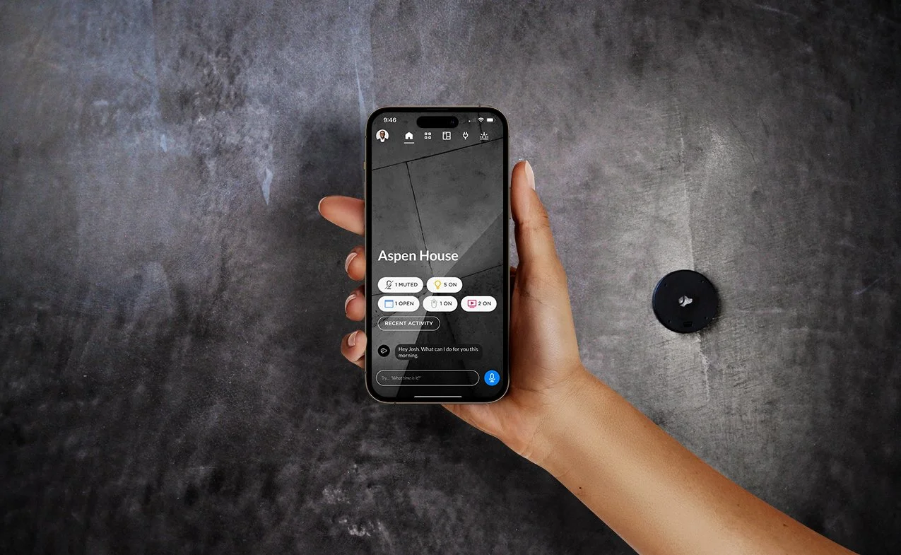Josh AI app held in one hand showing home status and a voice prompt, with a wall-mounted Josh Nano in the background.