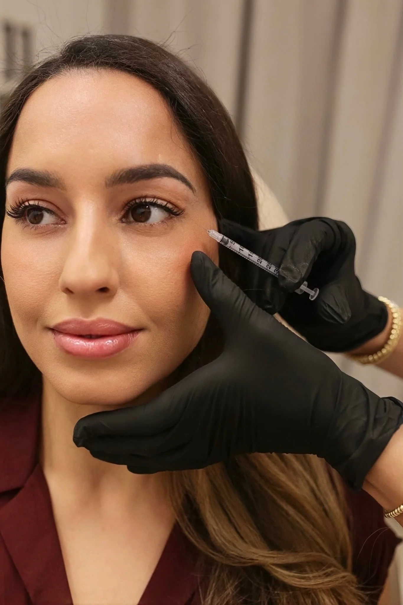 A person smiles while receiving a Botox injection near the outside corner of her eye from a healthcare professional in a cosmetic clinic.