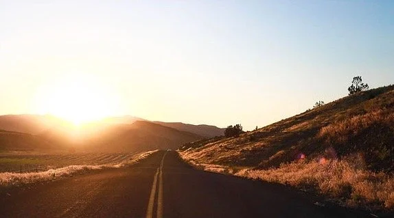 Open road leading toward the sunset over rolling hills with clear sky.