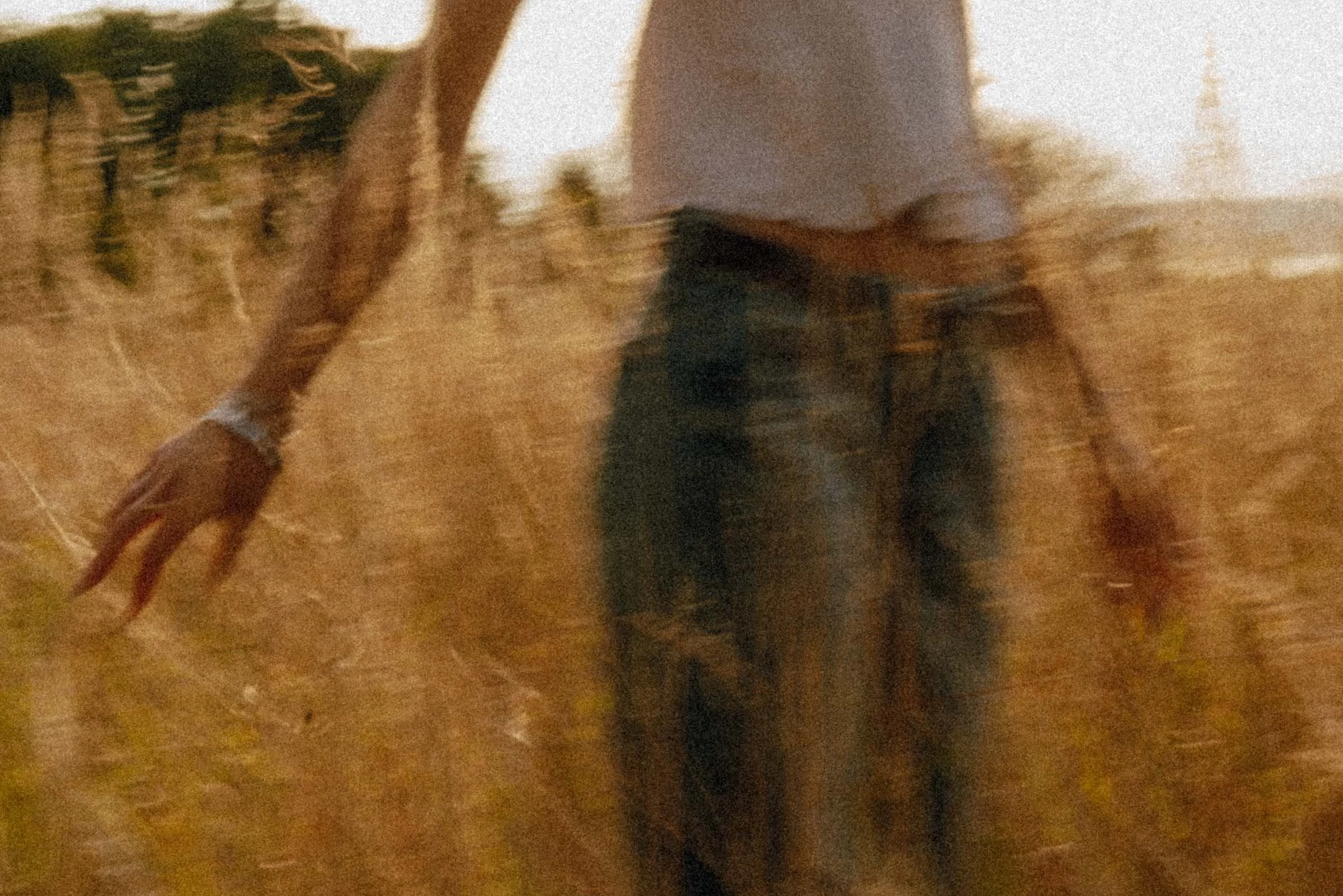 A person walking through a dry, grassy field at sunset, wearing a white shirt and dark pants, with other trees and foliage in the background.