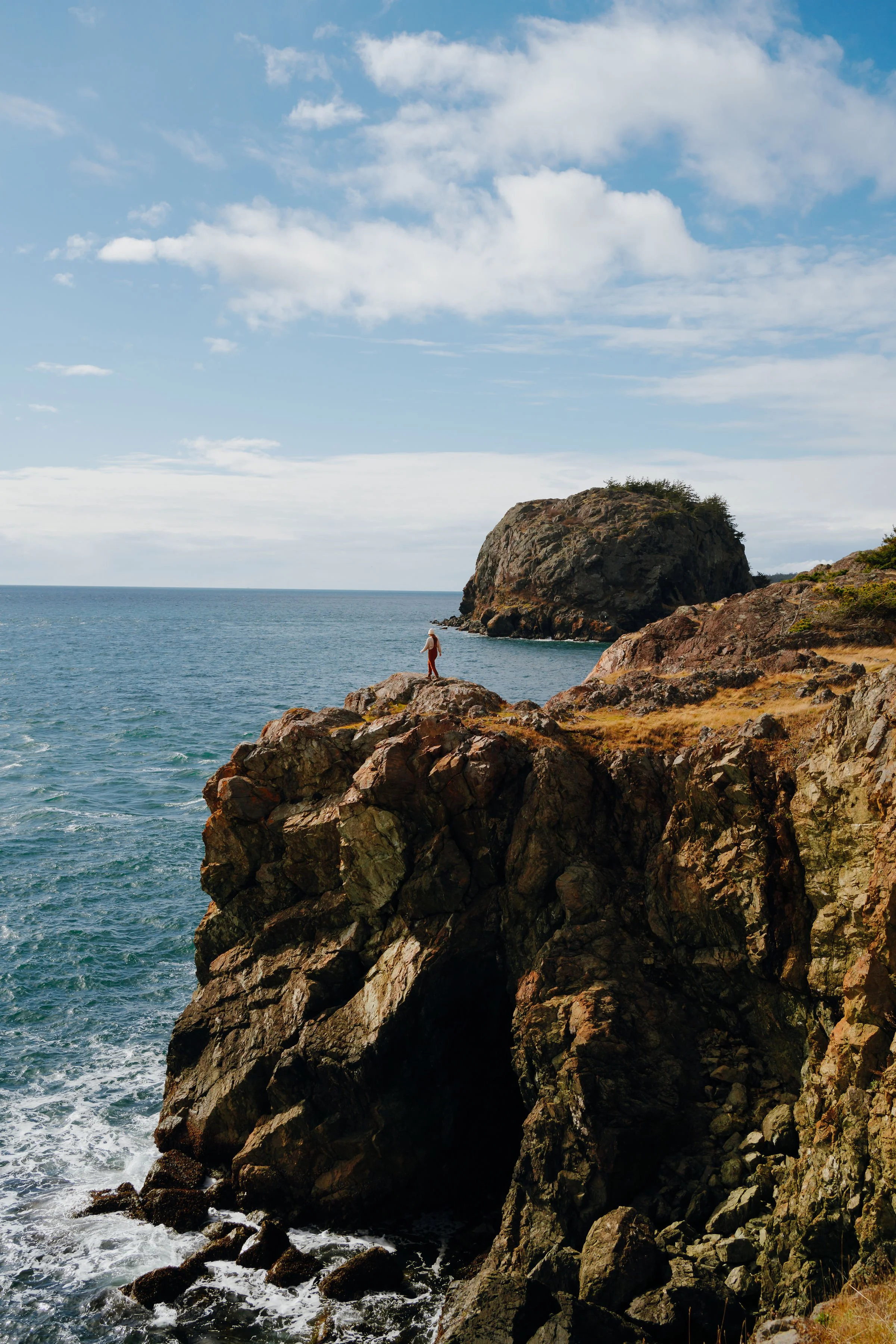 A person standing on a rocky cliff overlooking the ocean, with a large rock formation in the background and a partly cloudy sky.
