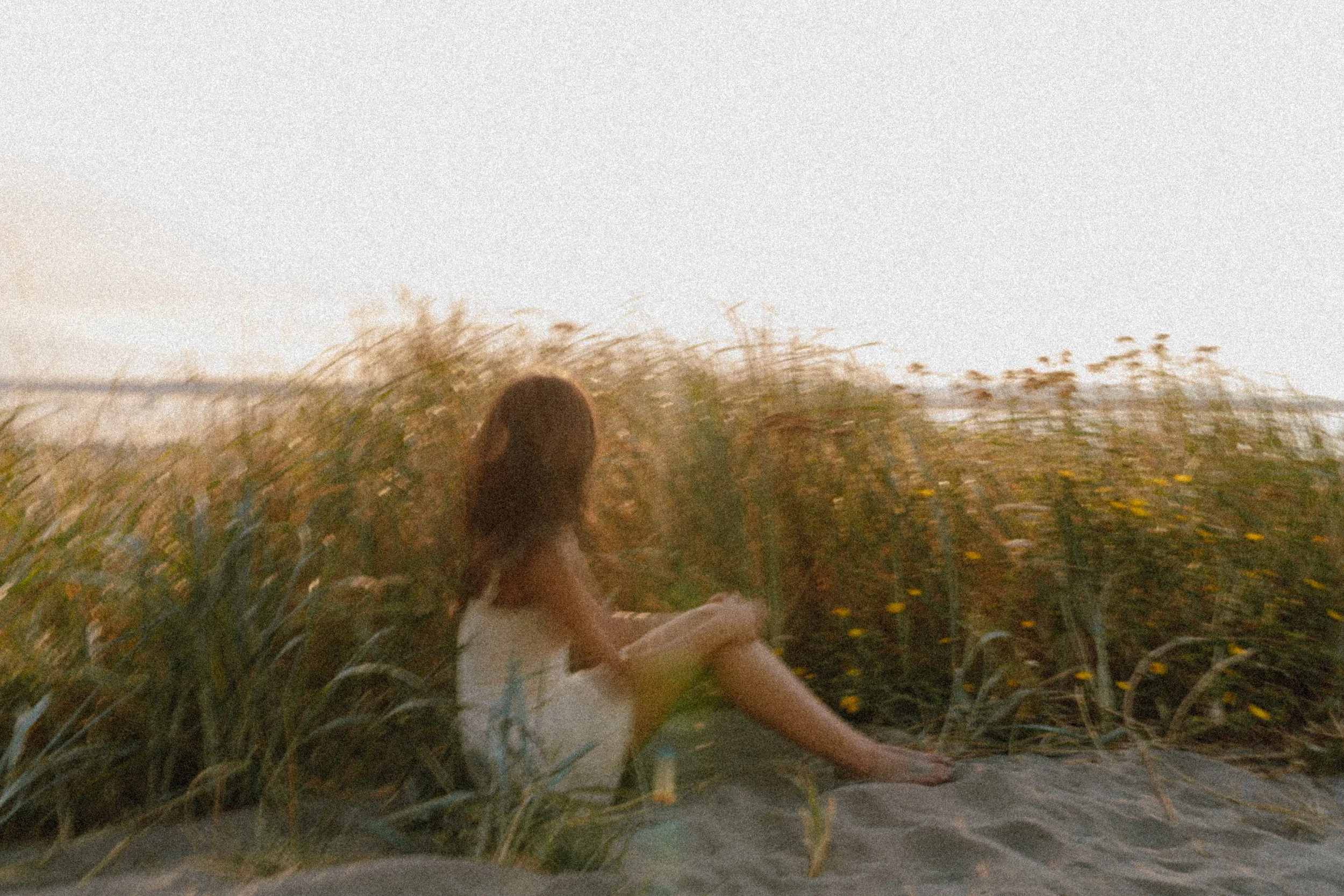 A woman lying in a field of tall grass during sunset or sunrise, with her arms relaxed, wearing a black top, black shorts, and jewelry.