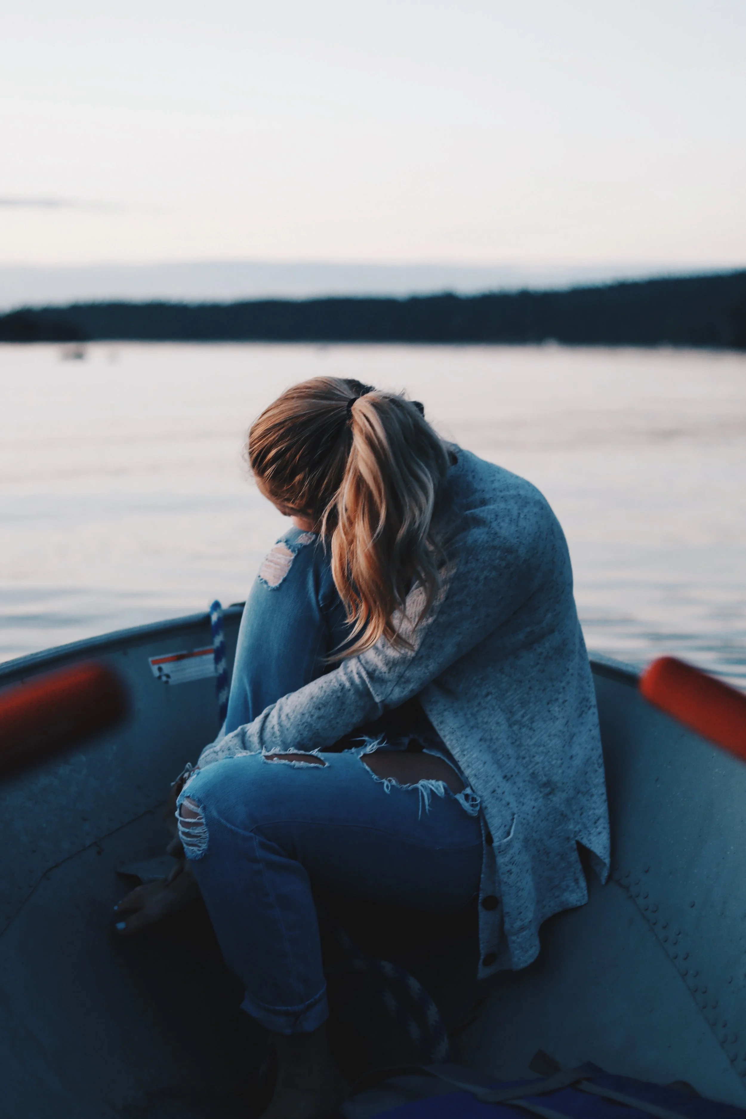 A woman sits in a boat on a calm lake during sunset, hugging her knees and looking downward, with a forested shoreline in the background.