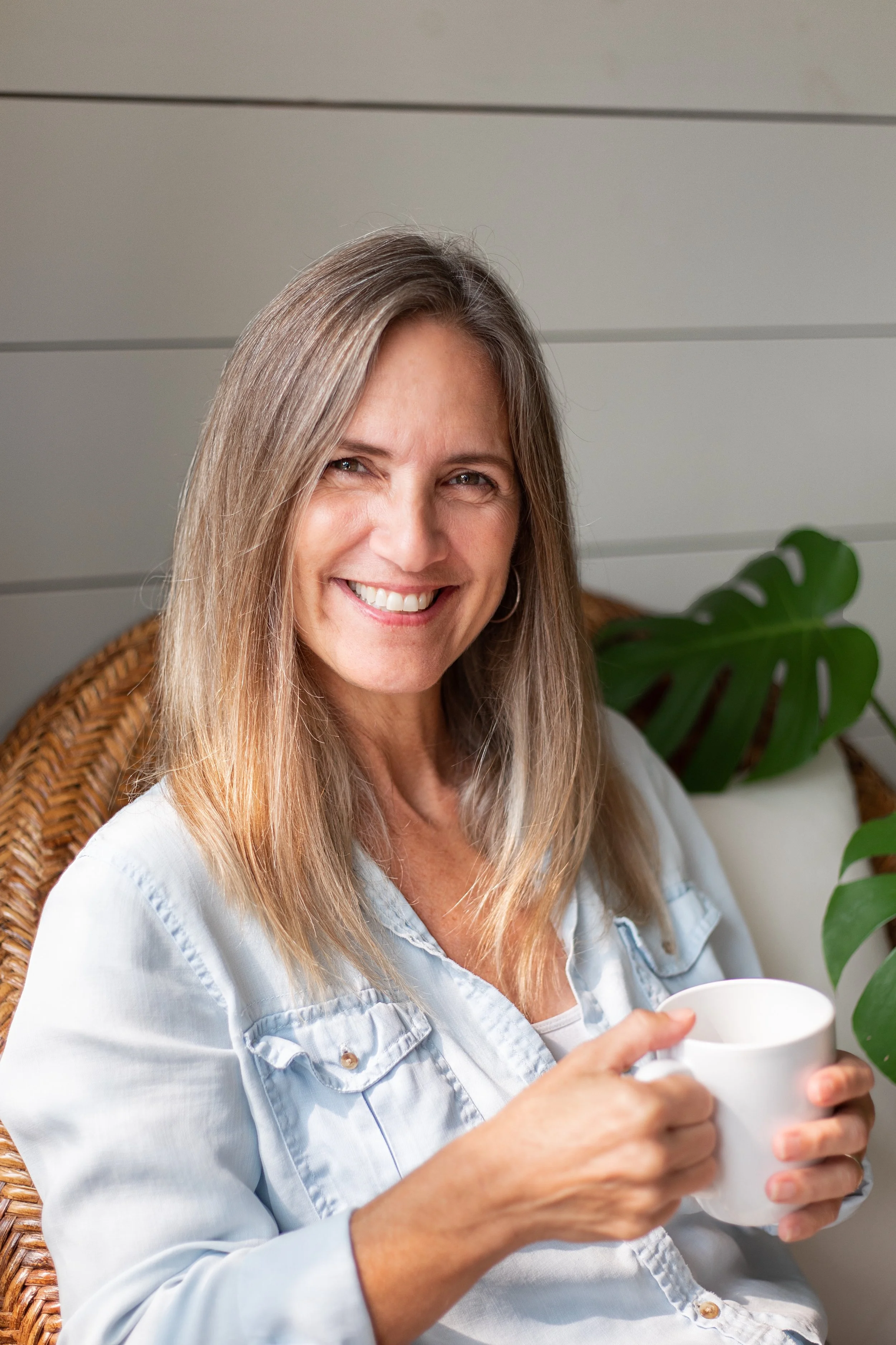 A woman with long blonde hair smiling and holding a white mug while sitting on a wicker chair with a green plant nearby.