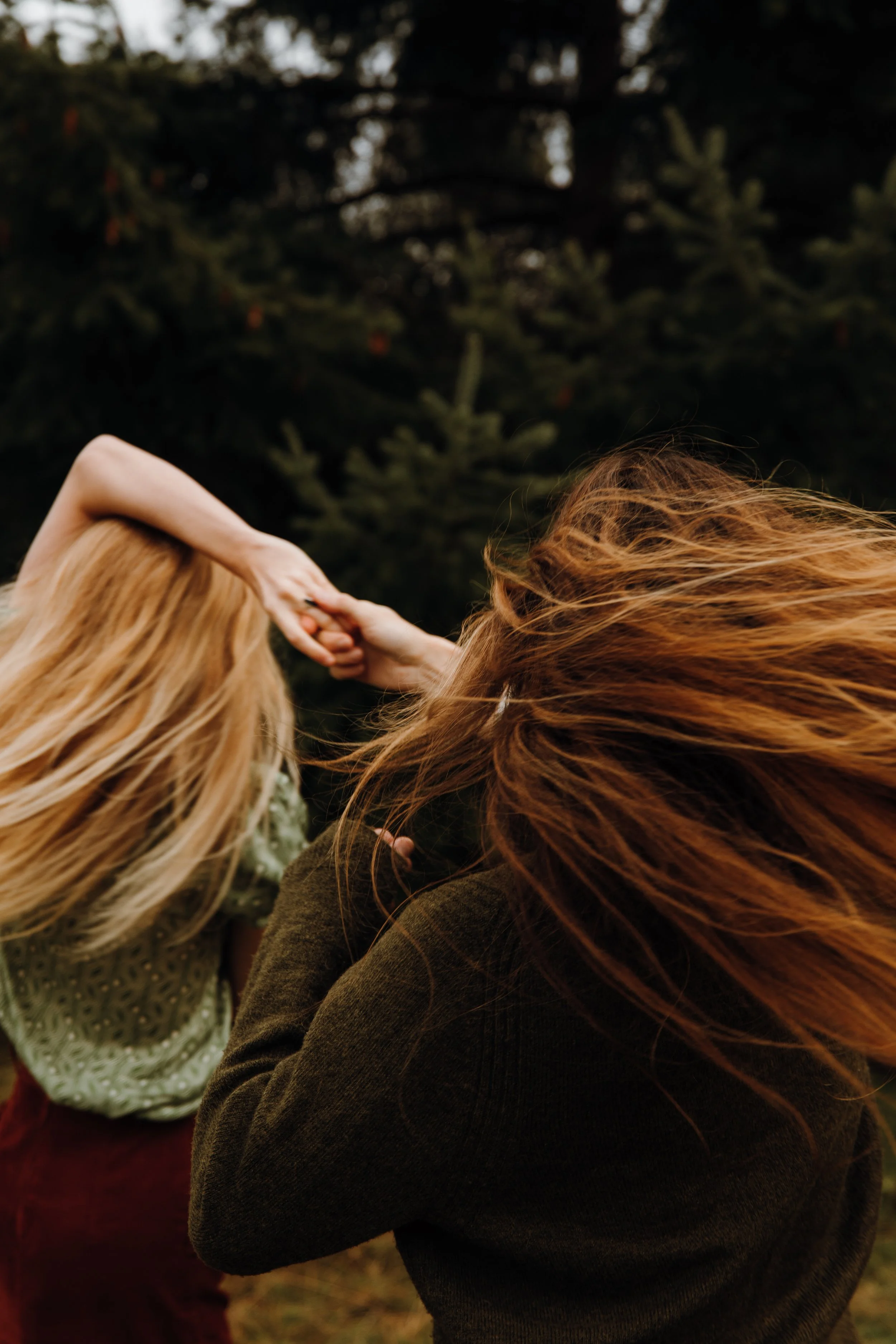 Two women with long red hair holding hands outdoors near tall trees.