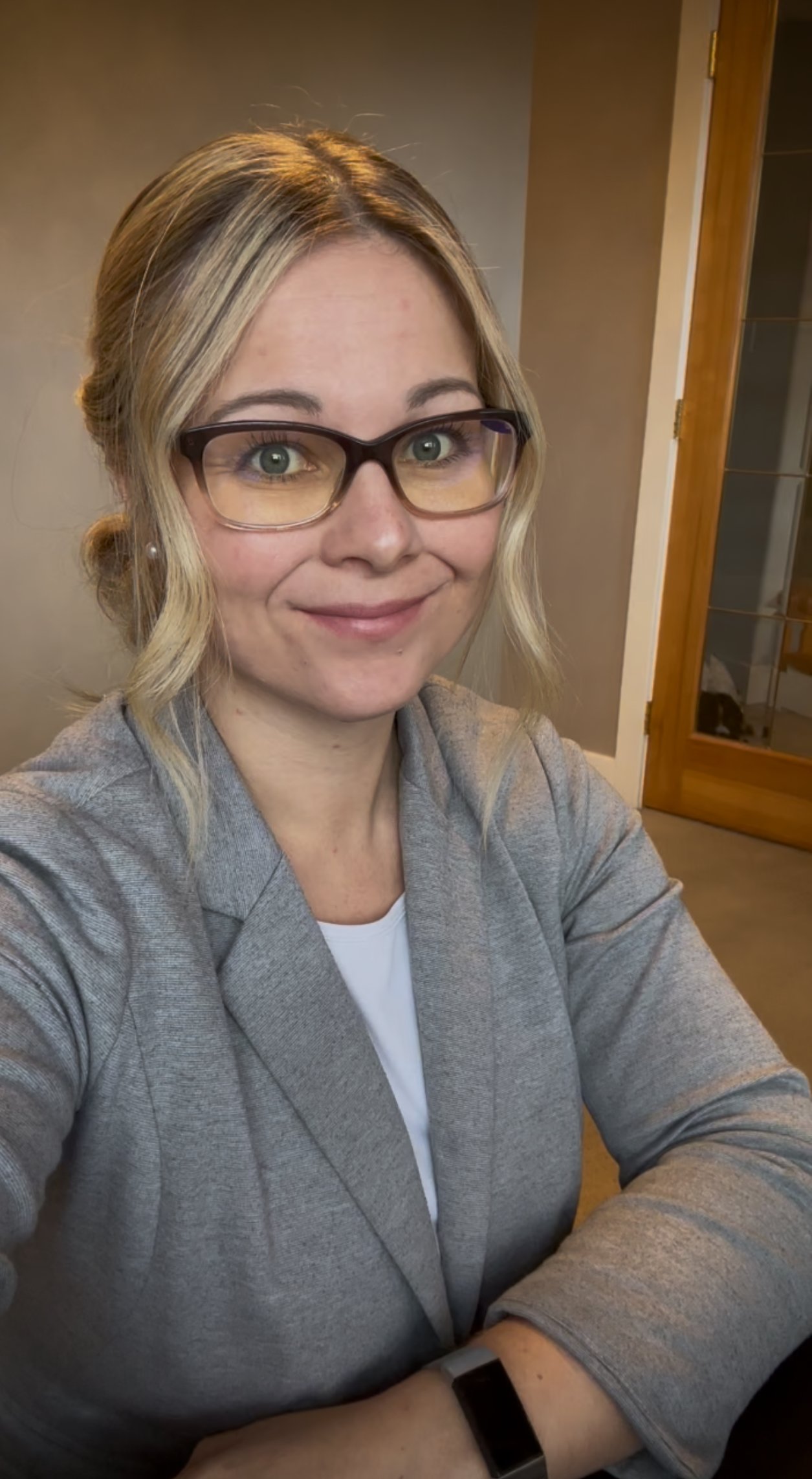 A woman with shoulder-length blond hair, wearing glasses, a gray blazer, and a white shirt, sitting at a table with a smile, in a room with a beige wall and a wooden door with glass panels.