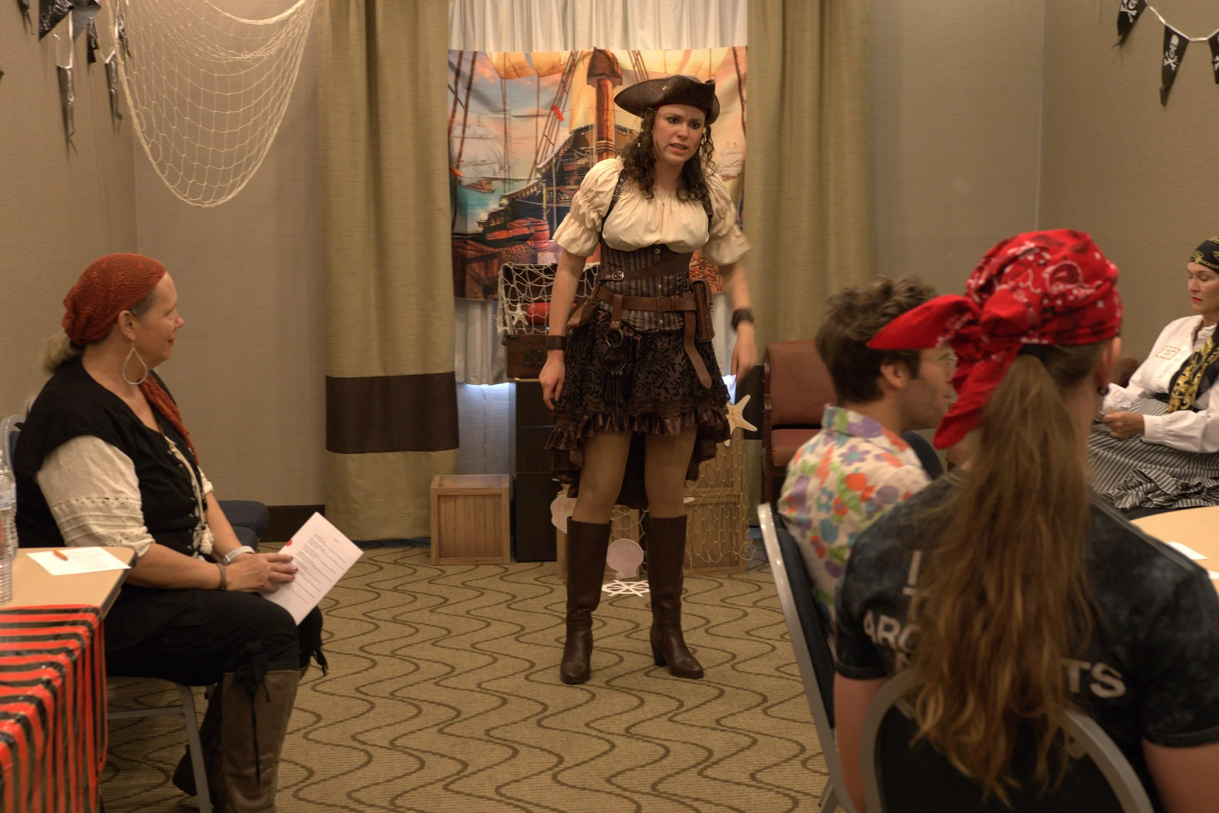 A modern building with wooden vertical slats on its front face, large glass window, and a gray roof, set against a mountainous landscape with dry grass and small trees in the foreground.