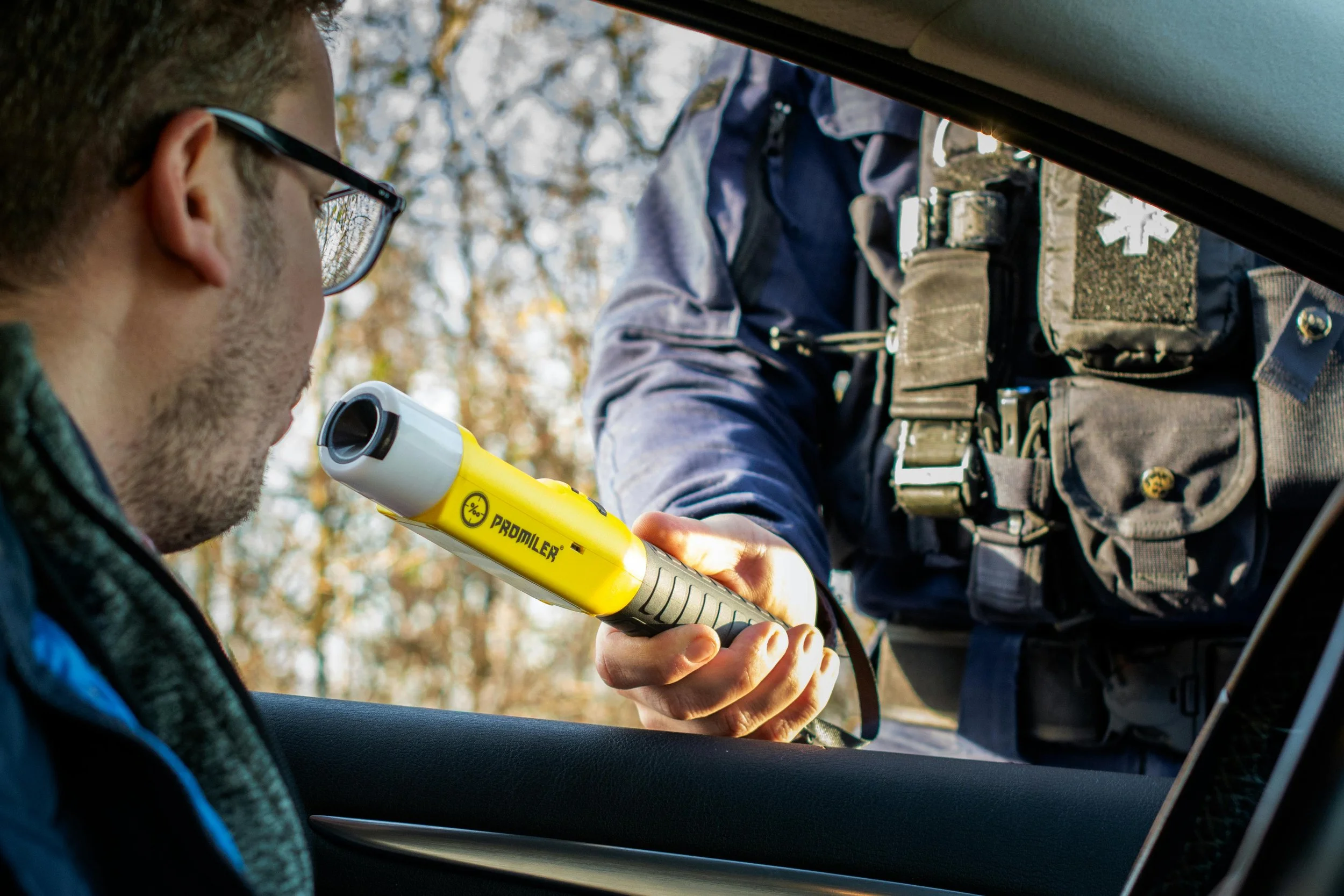 A police officer is holding a yellow breathalyzer near a man inside a vehicle, conducting a sobriety test outdoors with trees in the background.