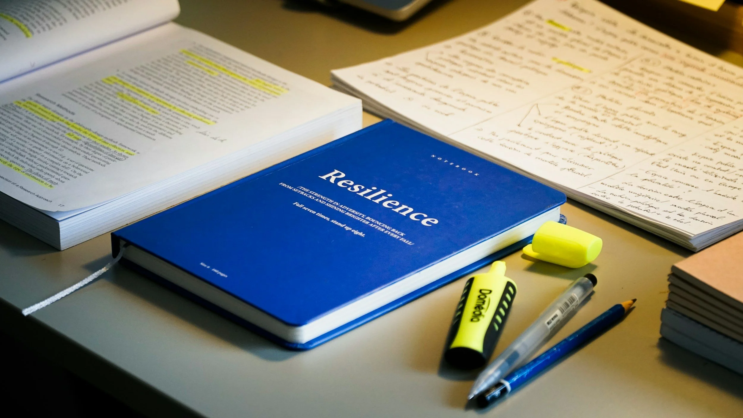 A desk with an open textbook, a stack of handwritten notes, and a blue book titled 'Resilience' surrounded by a yellow highlighter, a yellow and black pen, and a clear pen.