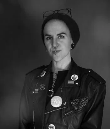 A woman with short dark hair wearing a dark blazer with multiple buttons and pins, glasses resting on her head, and earrings.