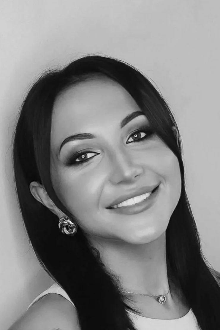 A woman with long dark hair, smiling, wearing earrings and a necklace, in black and white photography.