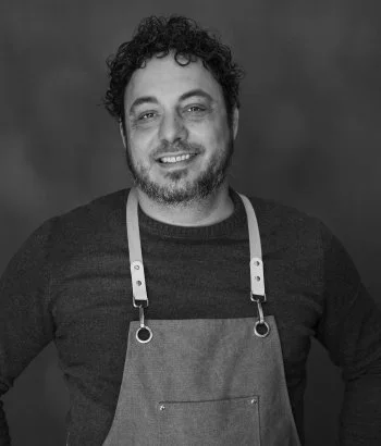 Black-and-white headshot of Paul Pugliese, Millheads Barbershop owner and barber-stylist, shown wearing an apron with dark hair in his mid-40s.
