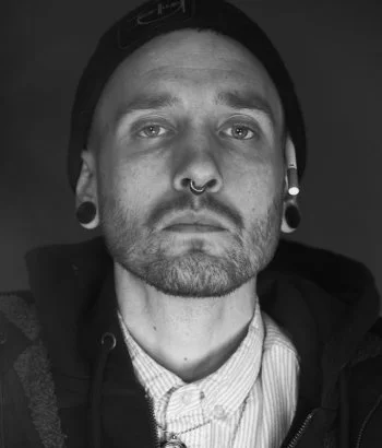 Black-and-white portrait of Daniel Wildermuth, Millheads barber and stylist, with multiple piercings, wearing a beanie, a white collared shirt, and a dark hooded sweatshirt, shown with a serious expression.