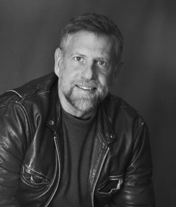 Black-and-white portrait of Jamieson, a bearded Millheads barber, smiling in a leather jacket and dark shirt against a plain background.