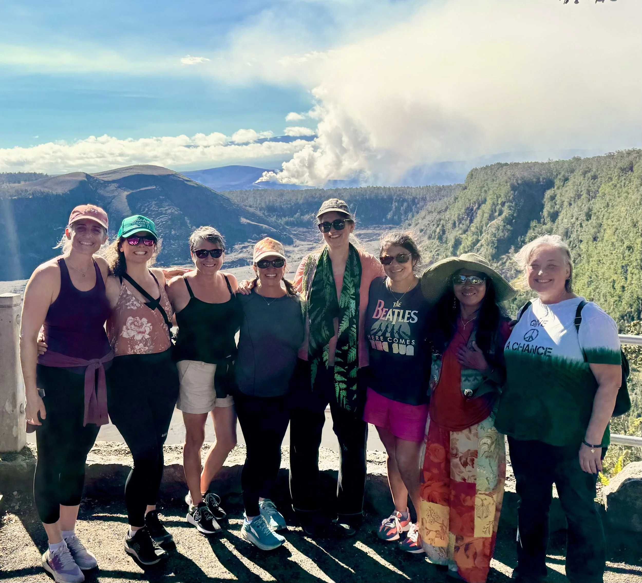 Group of eight women standing together outdoors with mountains and clouds in the background, smiling at the camera in Volcano National Park.