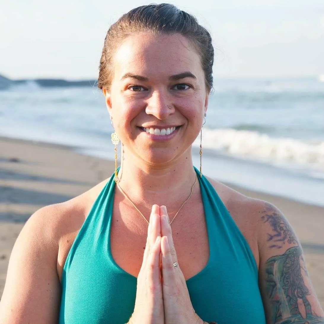 Woman smiling at the beach with hands in a prayer position, wearing a teal top, earrings, and a necklace, with ocean waves in the background.