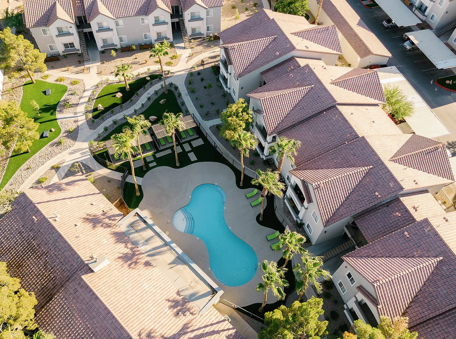 Aerial view of an apartment complex with a kidney-shaped swimming pool, surrounding lounge chairs, palm trees, green grassy areas, and a mix of pathways and parking spaces.