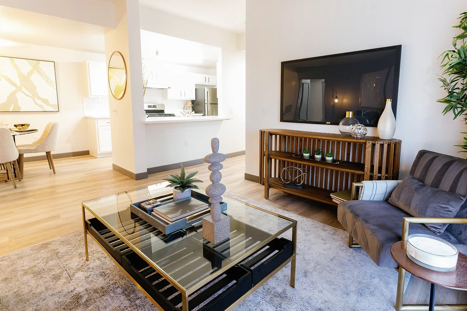 Living room with a gray couch, black and gold coffee table with decorative items, wooden TV stand with vases and plants, large flat-screen TV, small side table with a candle, and a large plant in the corner; open kitchen and dining area in background