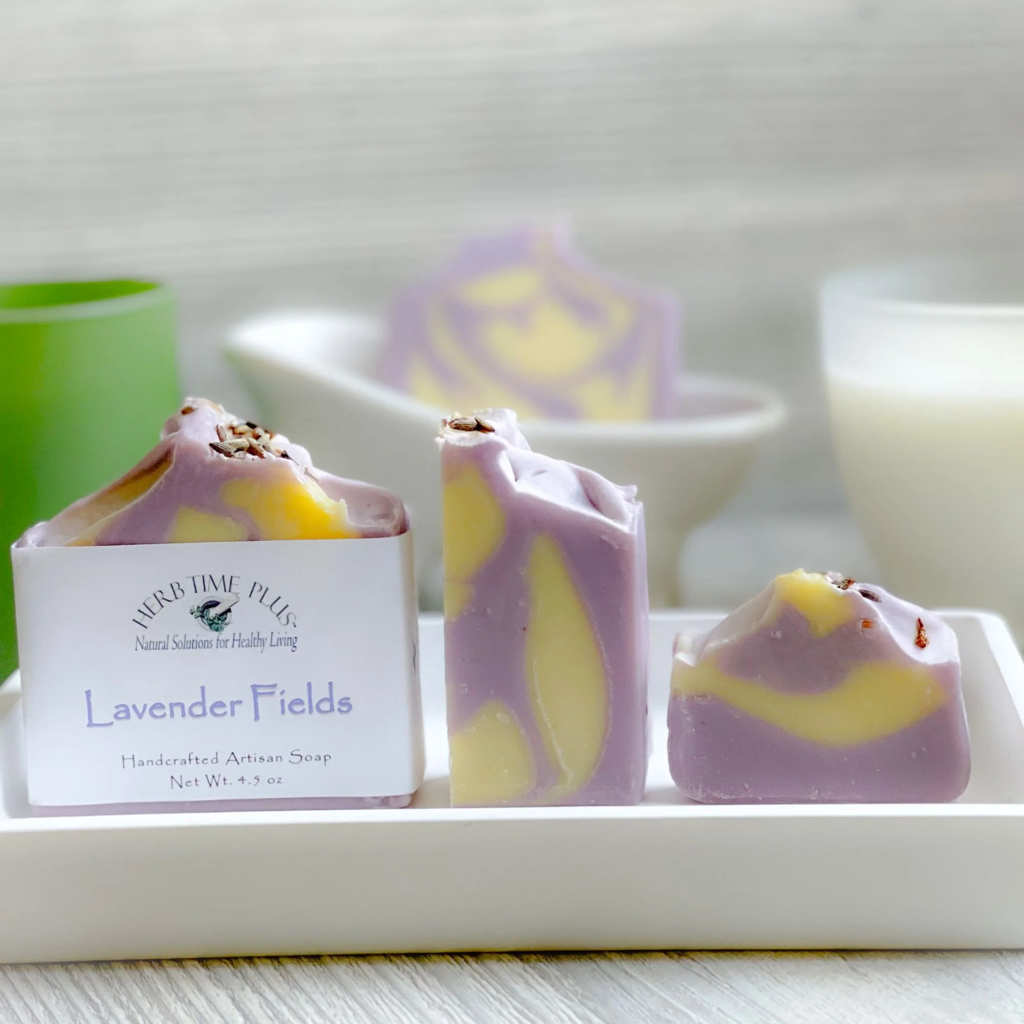lavender-fields-soap-bars.jpg: three different size soaps, one with a label on a soap tray with a soap in the background in a soap dish and candles on the side of it