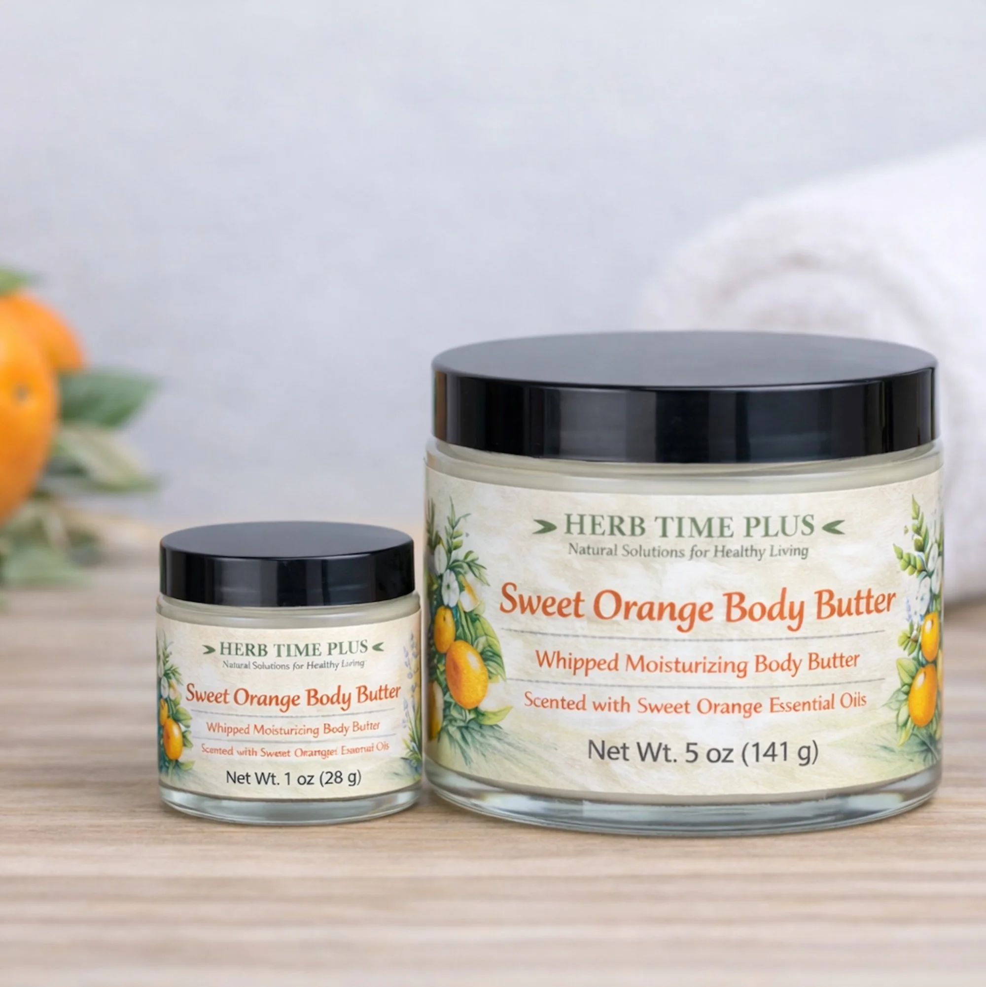 sweet-orange-body-butter.jpg: two jars of sweet orange body butter 1 oz and 5 oz on a table with a towel behind and an orange behind them