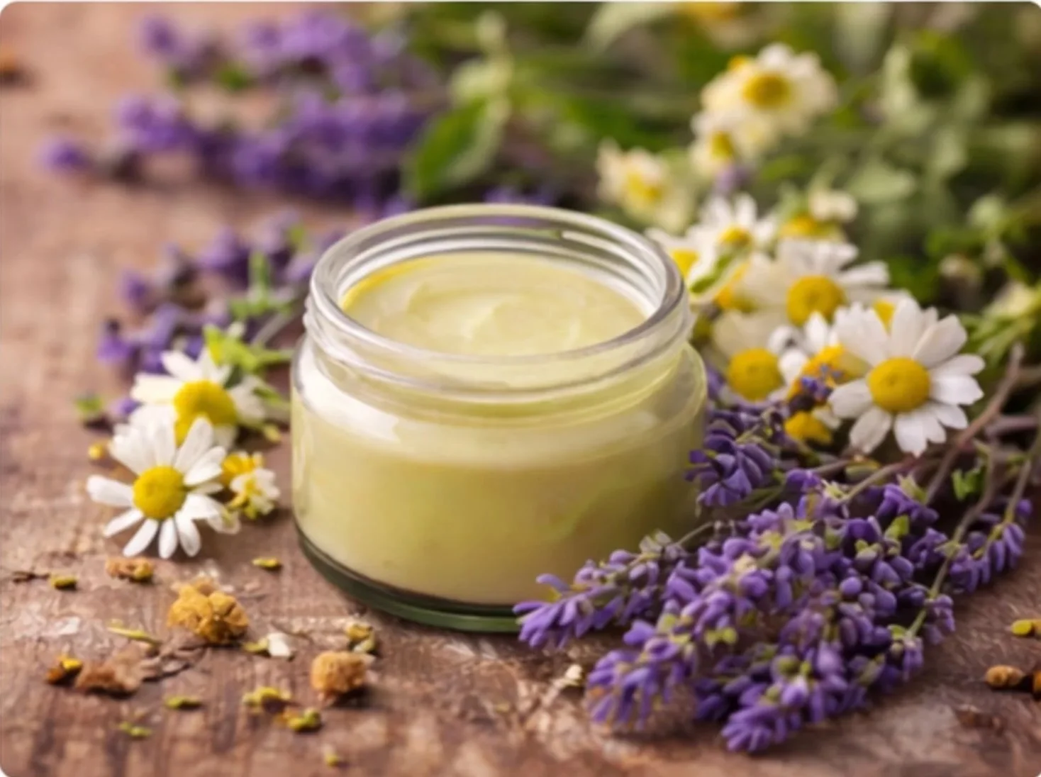 A small jar of light yellow balm surrounded by sprigs of lavender and chamomile flowers on a rustic wooden surface.