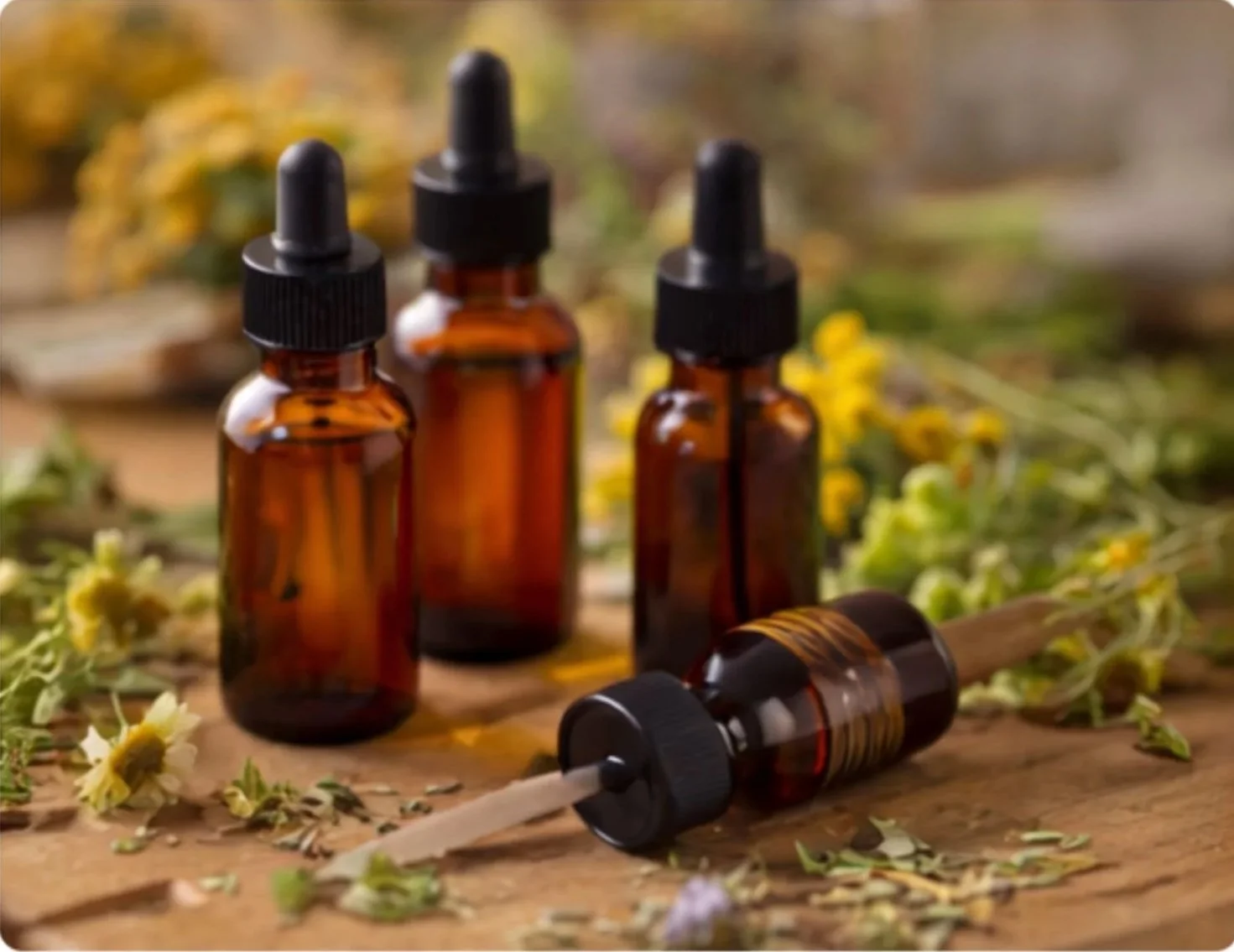 Four small amber glass dropper bottles with black caps and droppers, placed on a wooden surface surrounded by yellow and white flowers and green leaves.